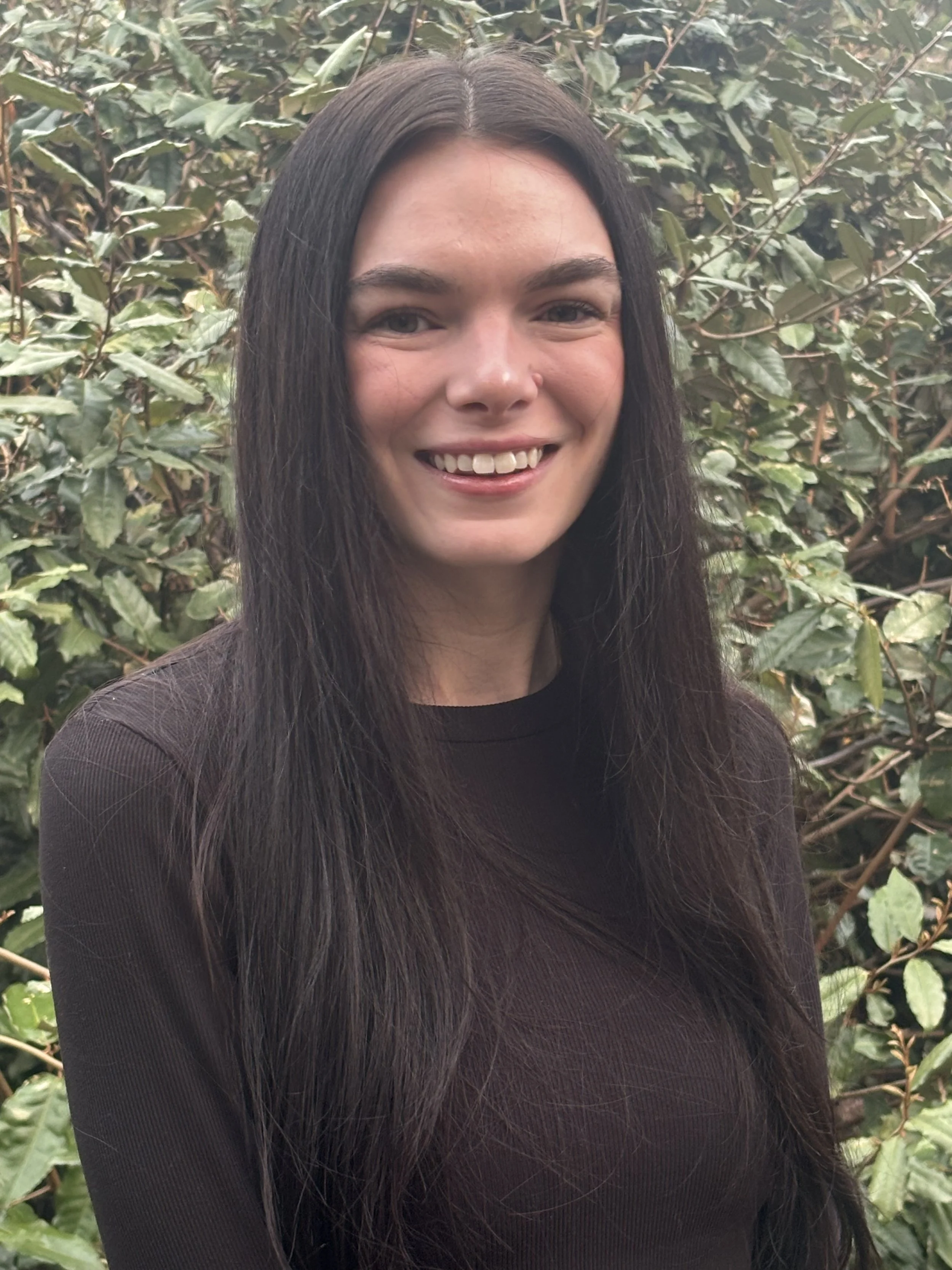 A young woman with long dark hair smiling outdoors in front of green leafy bushes.