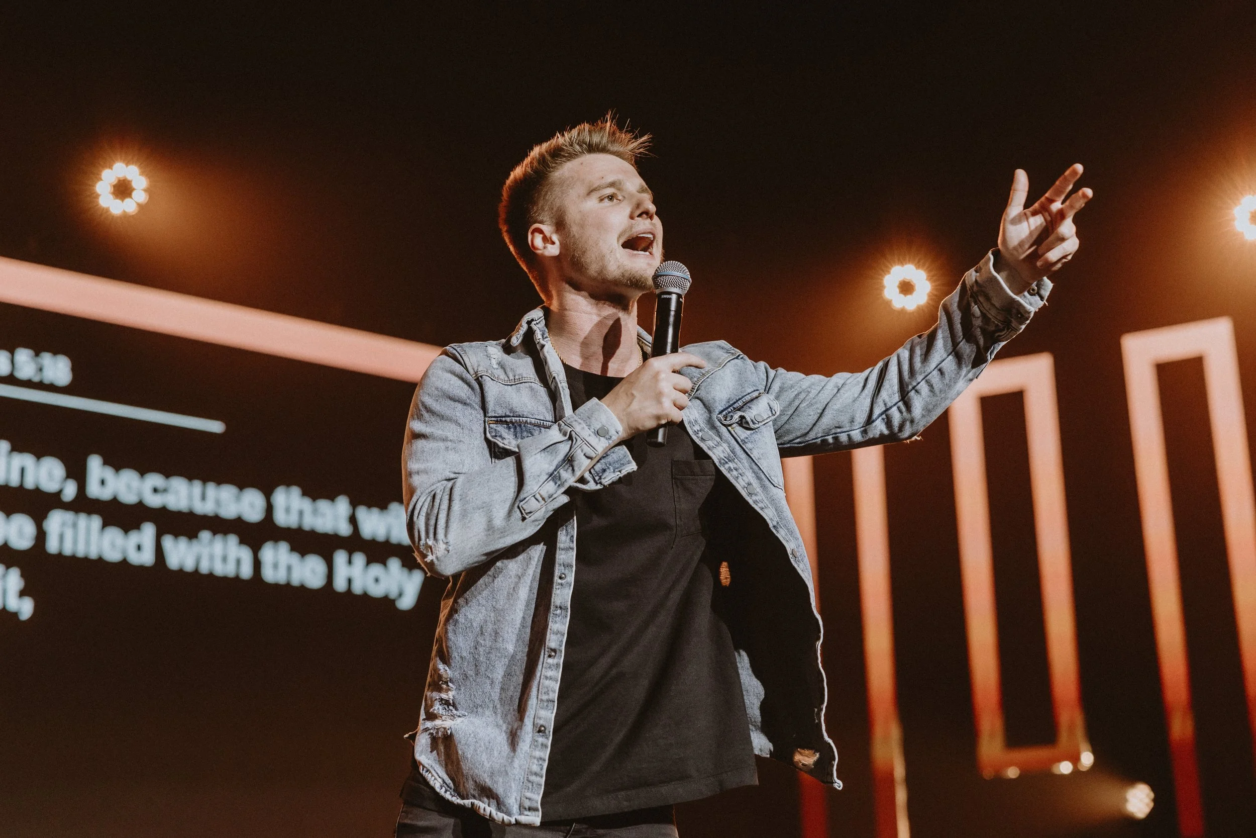 Young man speaking into a microphone on stage, wearing a denim jacket and black shirt, with warm-colored stage lights and a screen displaying text behind him.