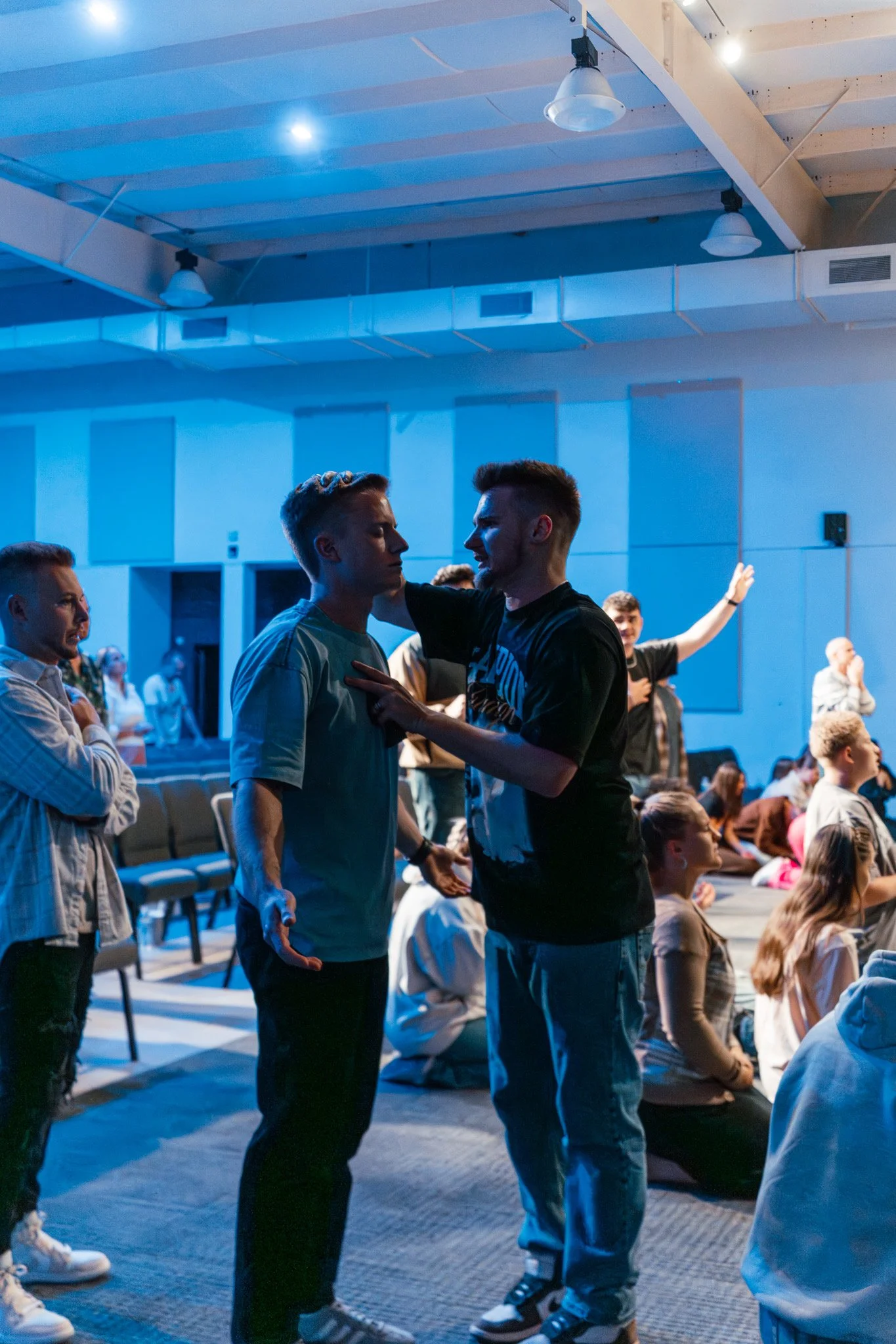 Two young men are engaged in a conversation, with one placing his hand on the other's chest. They are in a large indoor space with other people sitting and standing in the background. The ceiling has exposed beams and industrial lighting.