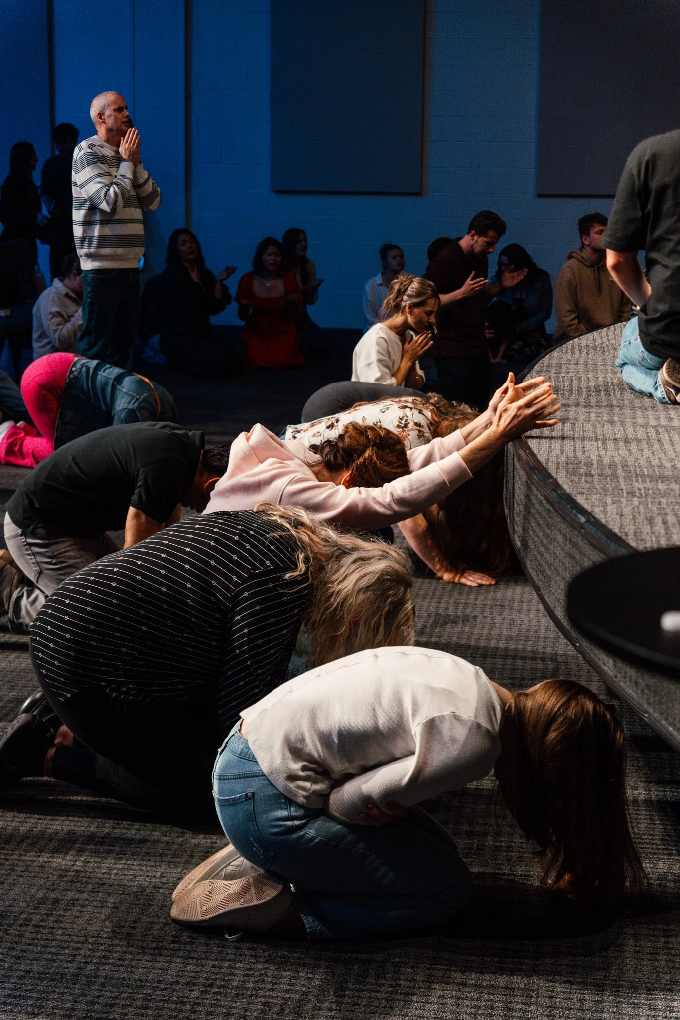 People kneeling and praying in a dimly lit room, some with hands clasped and heads bowed, others with hands raised or in prayerful pose.