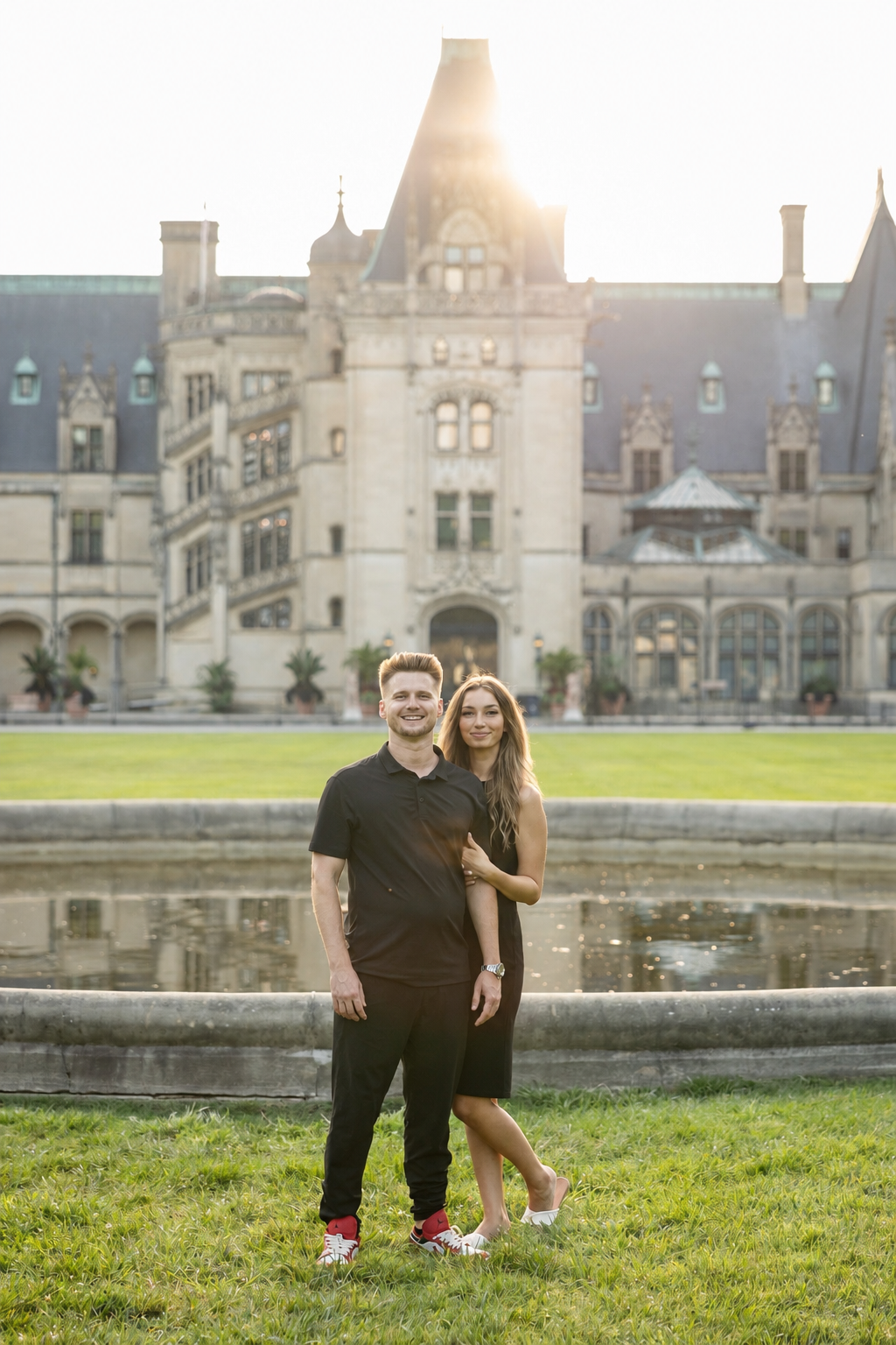 A young couple stands in front of a historic castle-like building with a pond in the foreground, sun shining in the background.