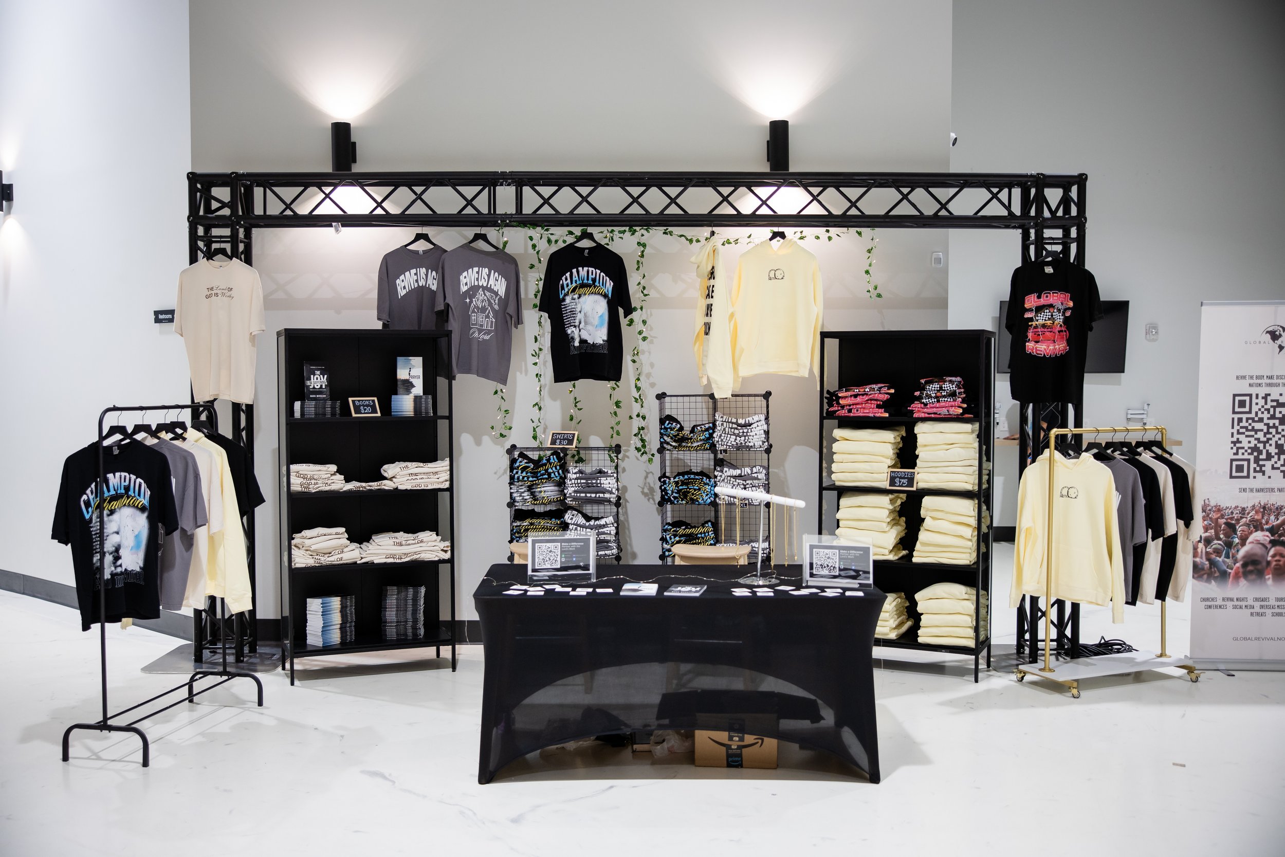 Display of clothing items at a retail booth, including t-shirts, hoodies, and folded sweatshirts with various designs, organized on black shelving units and racks, with signage and QR codes on a table in front.