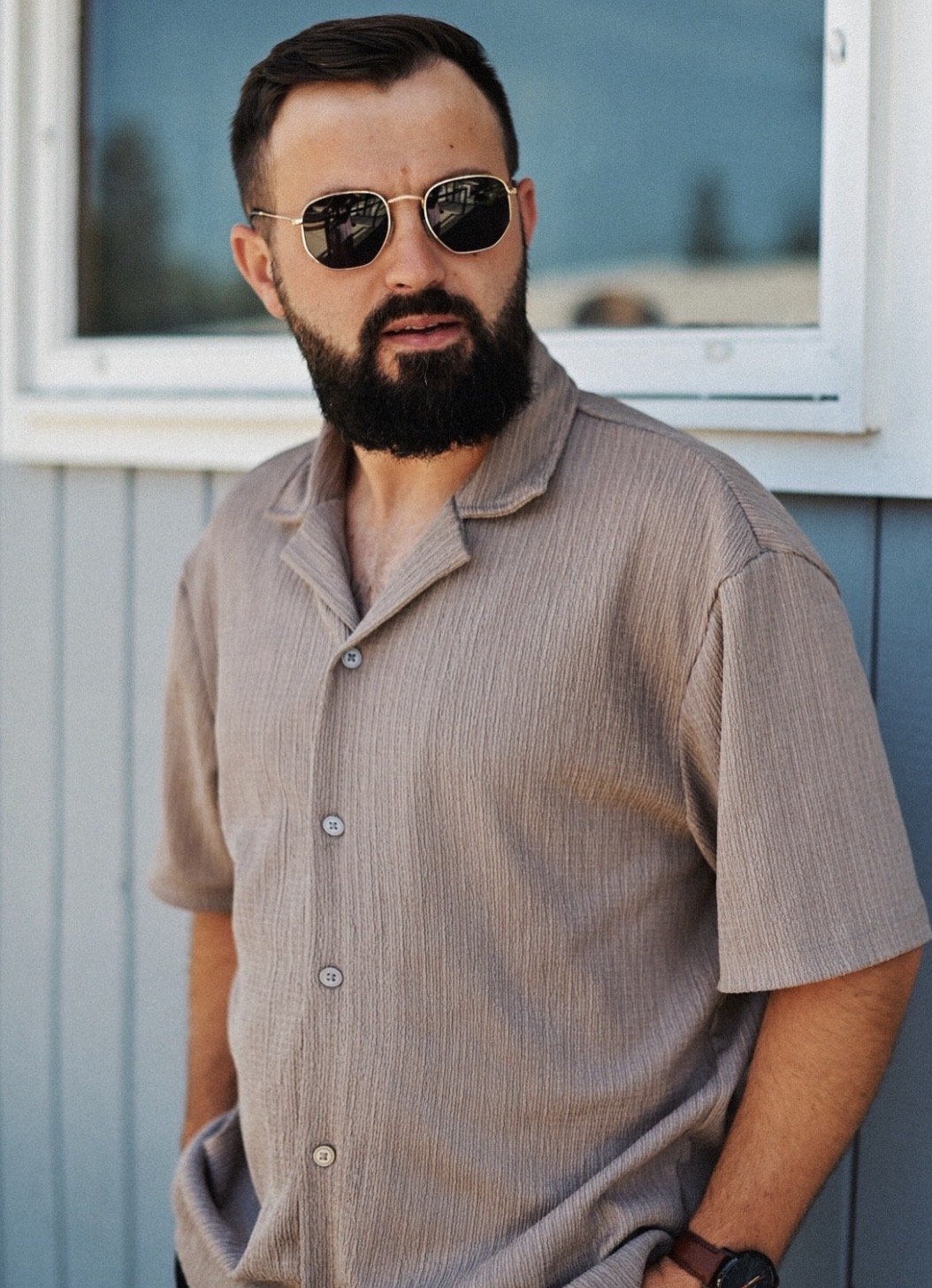 A man with short dark hair and a full beard wearing sunglasses and a beige short sleeve button-up shirt, standing outdoors near a building with light blue siding and a window.