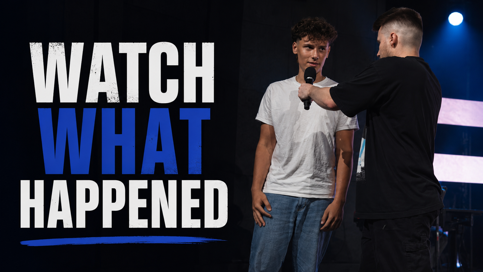 A young man with curly hair is being interviewed by an older man holding a microphone on stage, with a large sign displaying the text 'WATCH WHAT HAPPENED'.