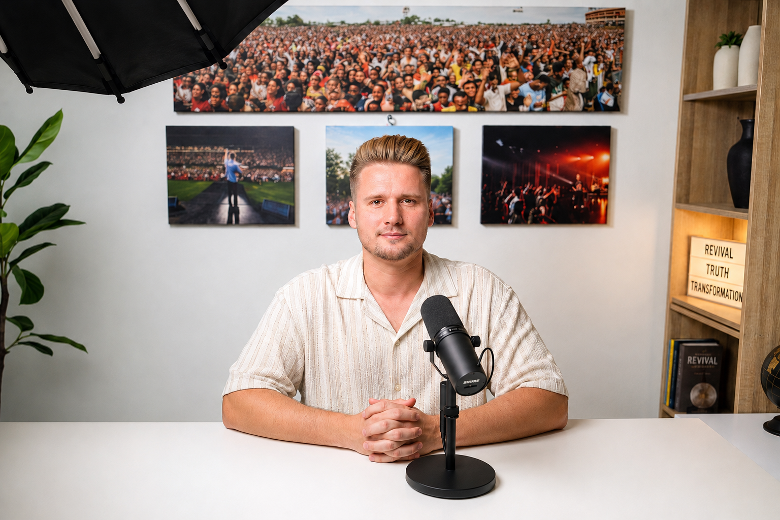 A man sitting at a desk with a microphone in front of him, in a room decorated with framed photos of large crowds, a bookcase with books, a potted plant, and a lightbox with words 'Revival,' 'Truth,' 'Transformation' written on it.