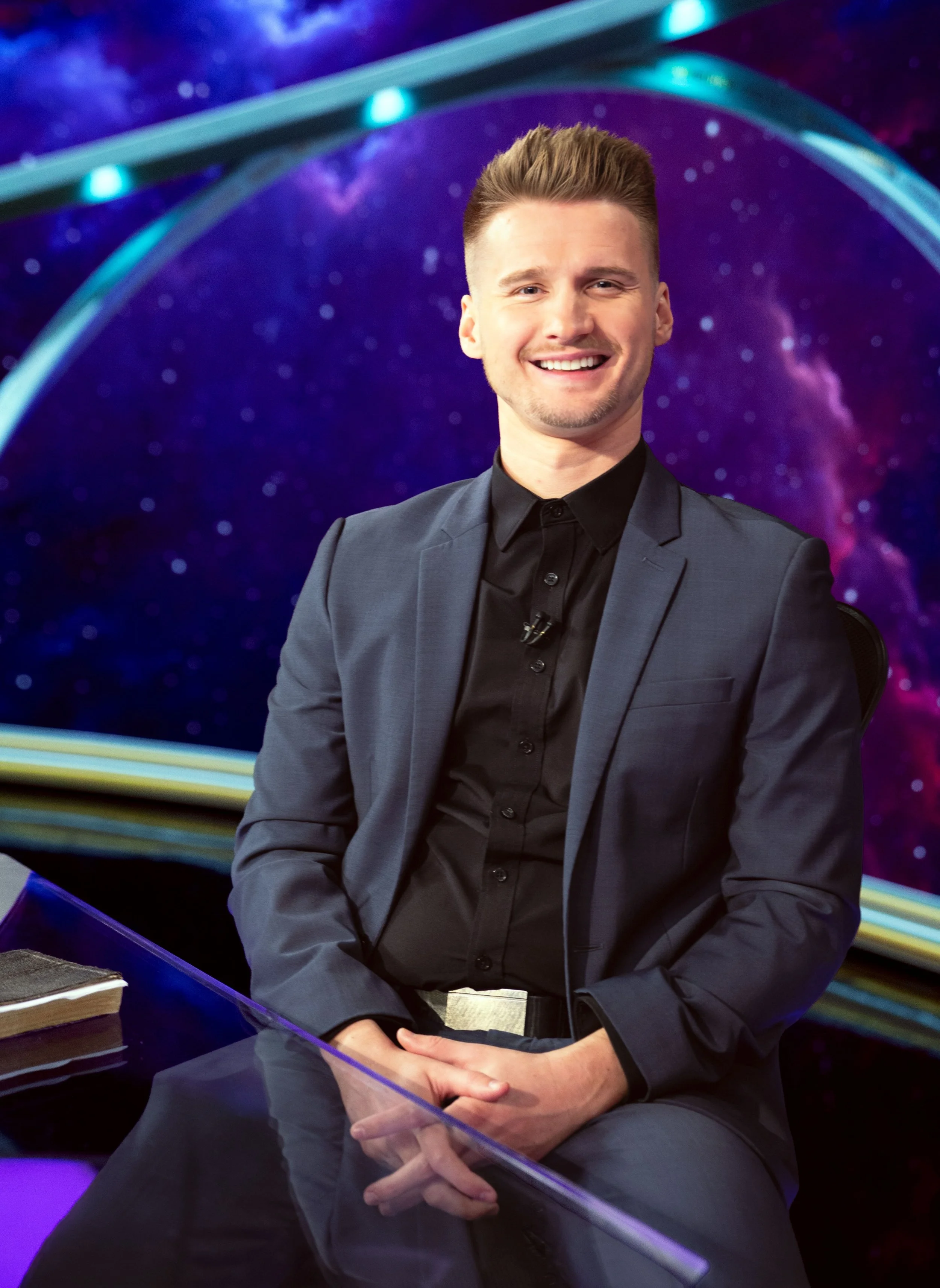 A young man smiling, wearing a gray suit and black shirt, sitting behind a transparent barrier with a colorful, space-themed background.