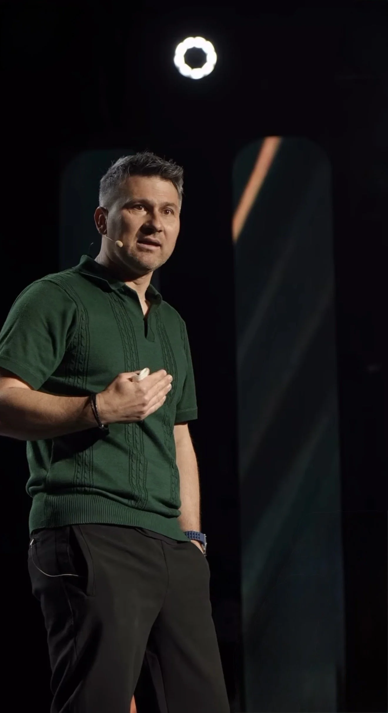 A man with short brown hair, wearing a green polo shirt and black pants, speaking on stage with a headset microphone.