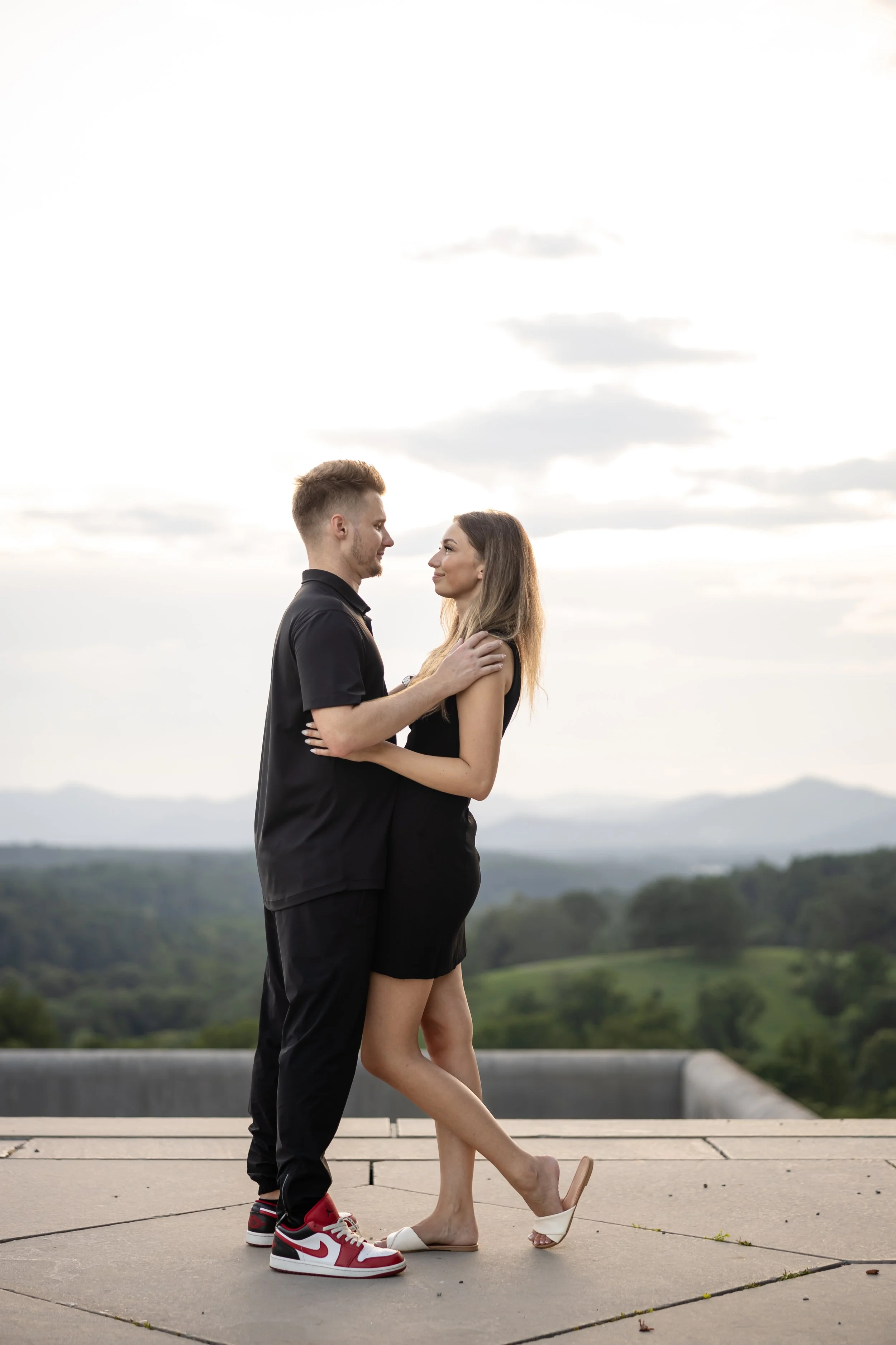 A man and woman are standing close to each other outdoors, facing each other with their foreheads almost touching. The man is wearing a black shirt, black pants, and red Nike sneakers, while the woman is dressed in a black dress and cream-colored high heels. They are standing on a paved surface with scenic green hills and sky with clouds in the background.