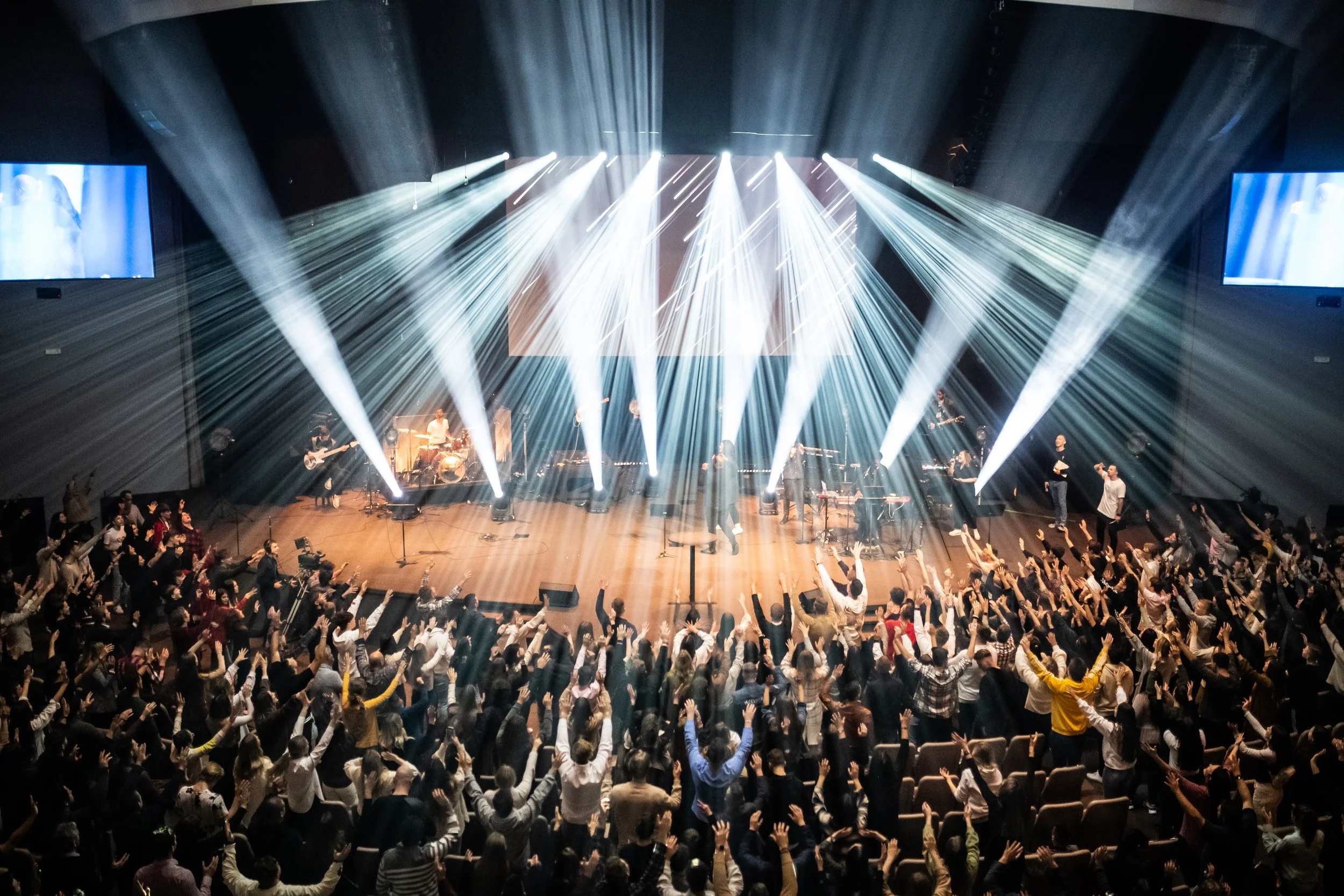 Large crowd in a theater or concert hall raising their hands towards a stage with performing musicians and bright stage lighting.