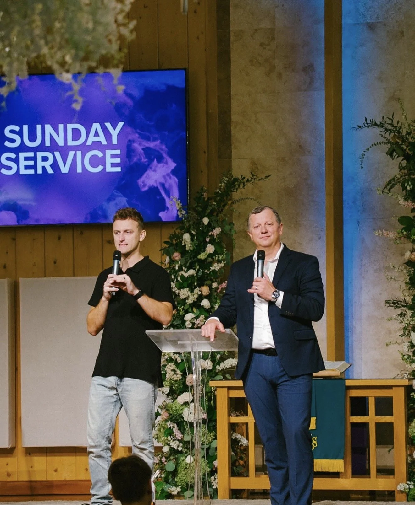 Two men speaking at a church service, one young man in a black shirt and jeans, and an older man in a suit, standing behind a clear pulpit, with a large screen in the background that reads 'Sunday Service' and decorated with flowers.