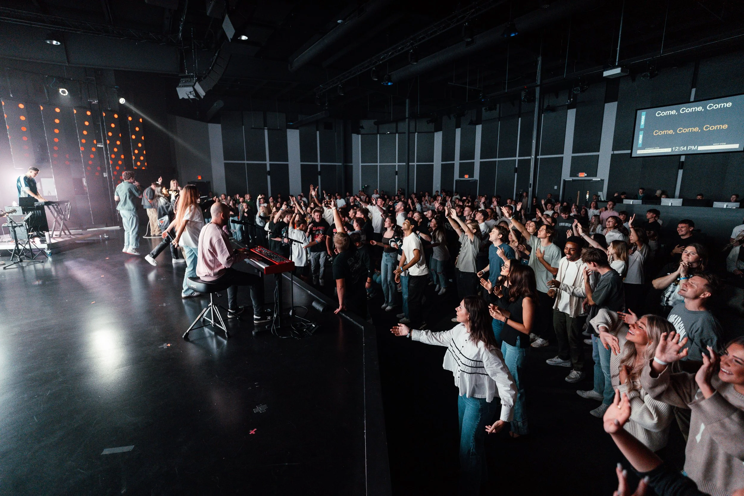 A crowd of people attending a concert or event in a dimly lit indoor venue with some standing and many raising their hands. A musician playing keys is on the stage on the left, and there are large screens and wall panels.