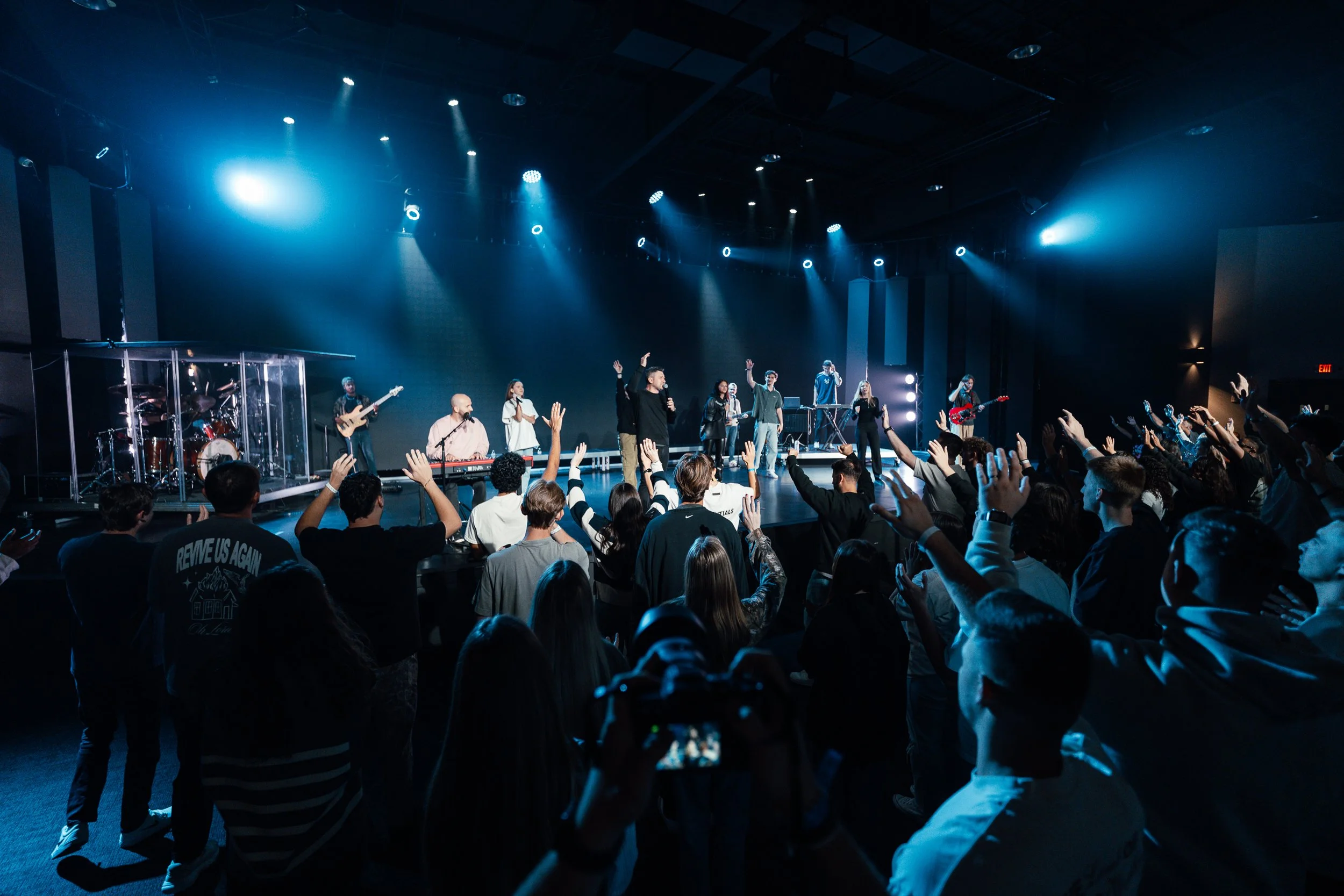 Crowd attending a live music concert with performers on stage, some with microphones, under blue stage lighting.