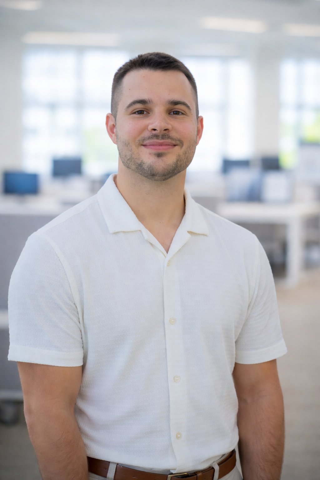 A young man with short dark hair and a trimmed beard, wearing a white short-sleeve button-up shirt, standing in a brightly lit office space with large windows and desks in the background.