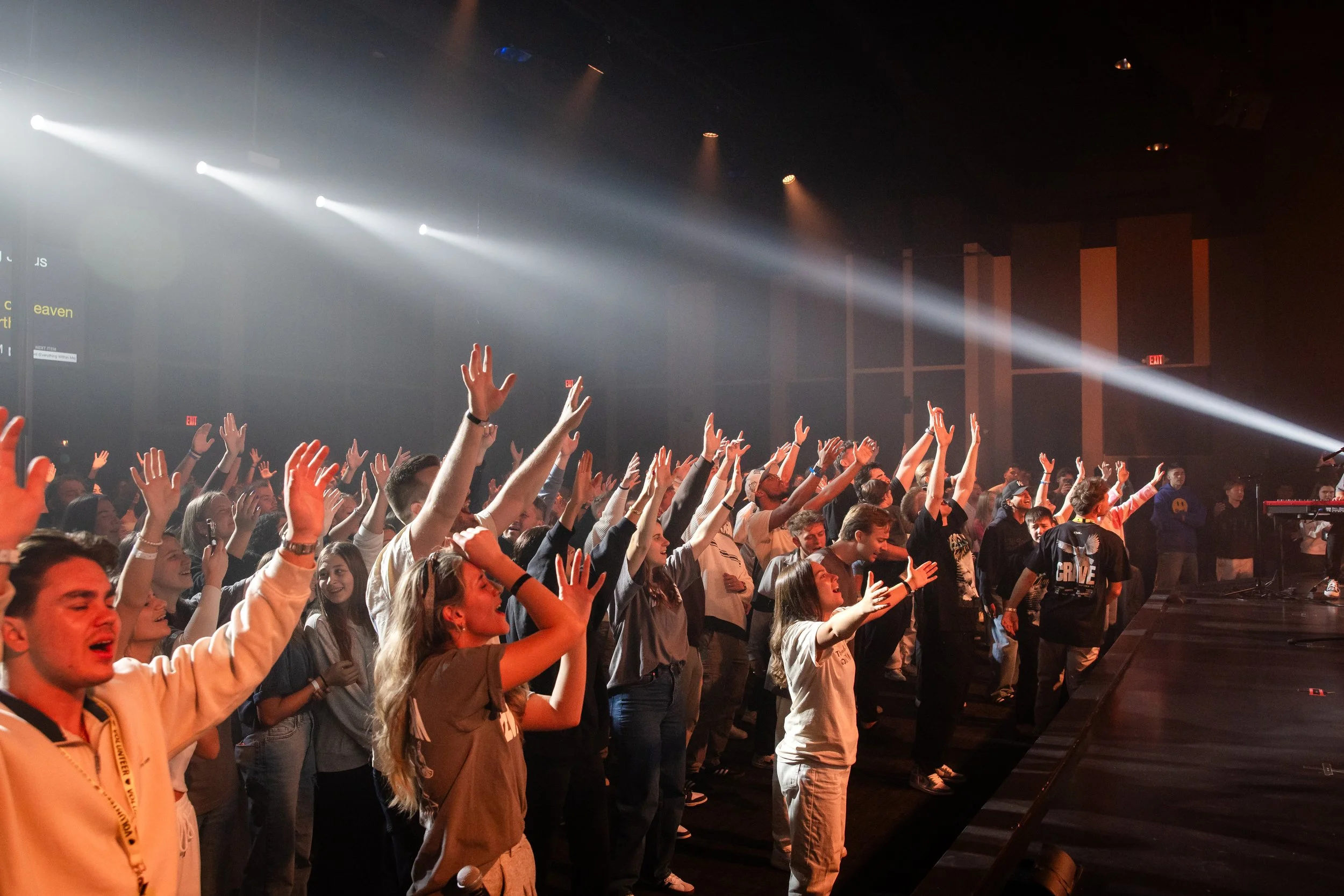 Crowd of young people at a concert with hands raised, stage lights illuminating the audience, and a dark concert hall background.