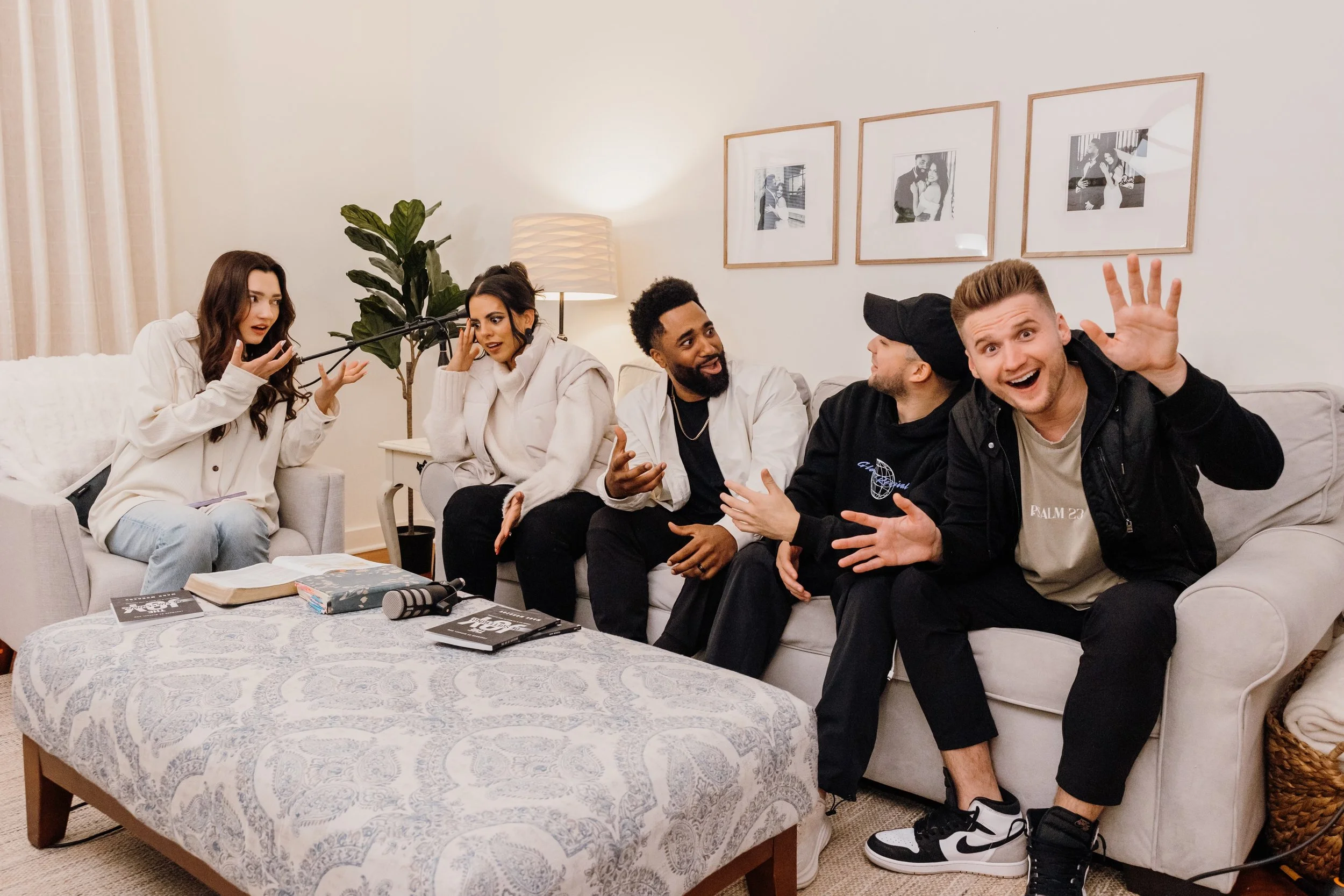 Five friends seated on a sofa in a living room, engaged in conversation and laughter, with a round coffee table in front, and framed photos on the wall behind them.