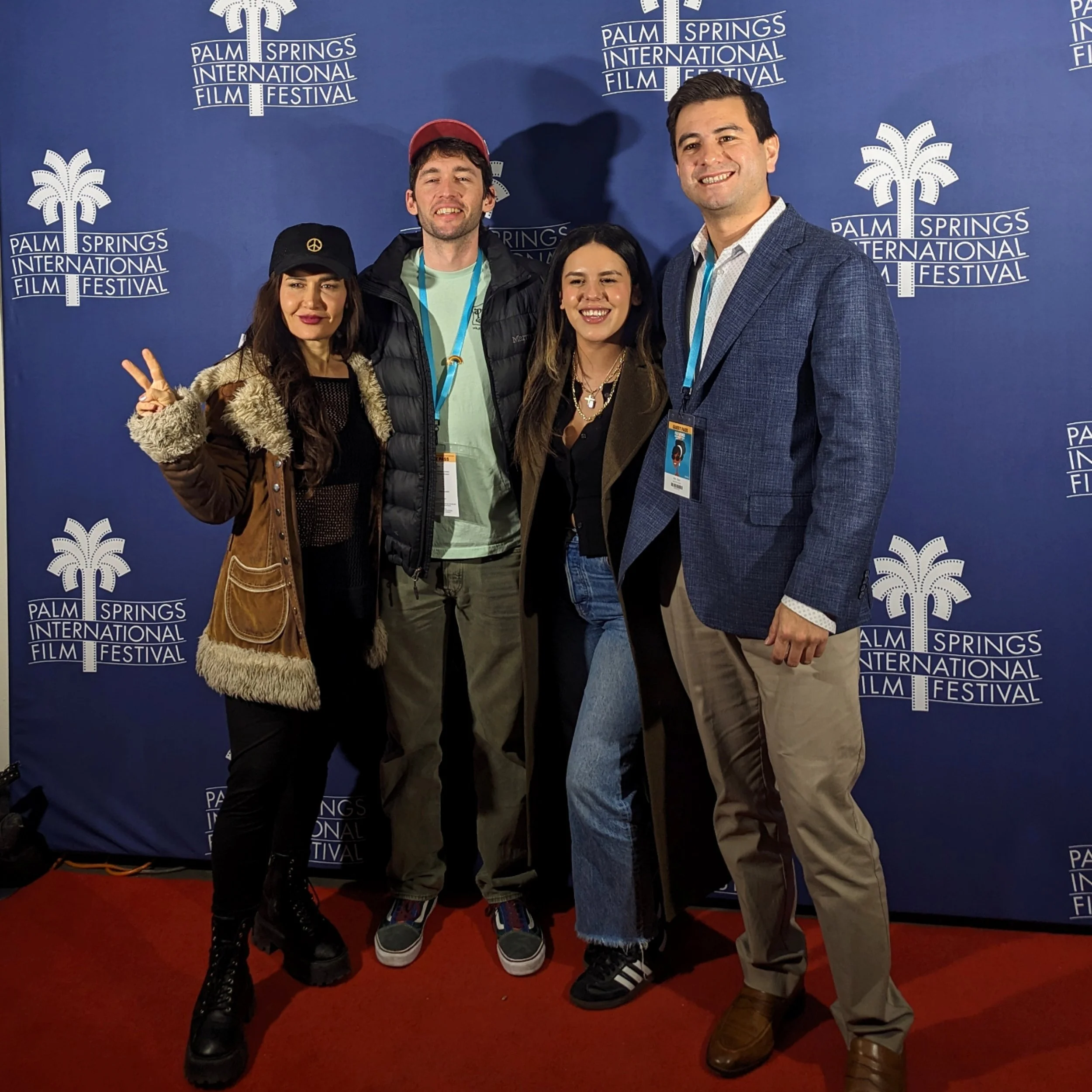 Four people standing in front of a blue backdrop with the Palm Springs International Film Festival logo. The group includes two women and two men, dressed casually. One woman is making a peace sign with her hand. Everyone is smiling.