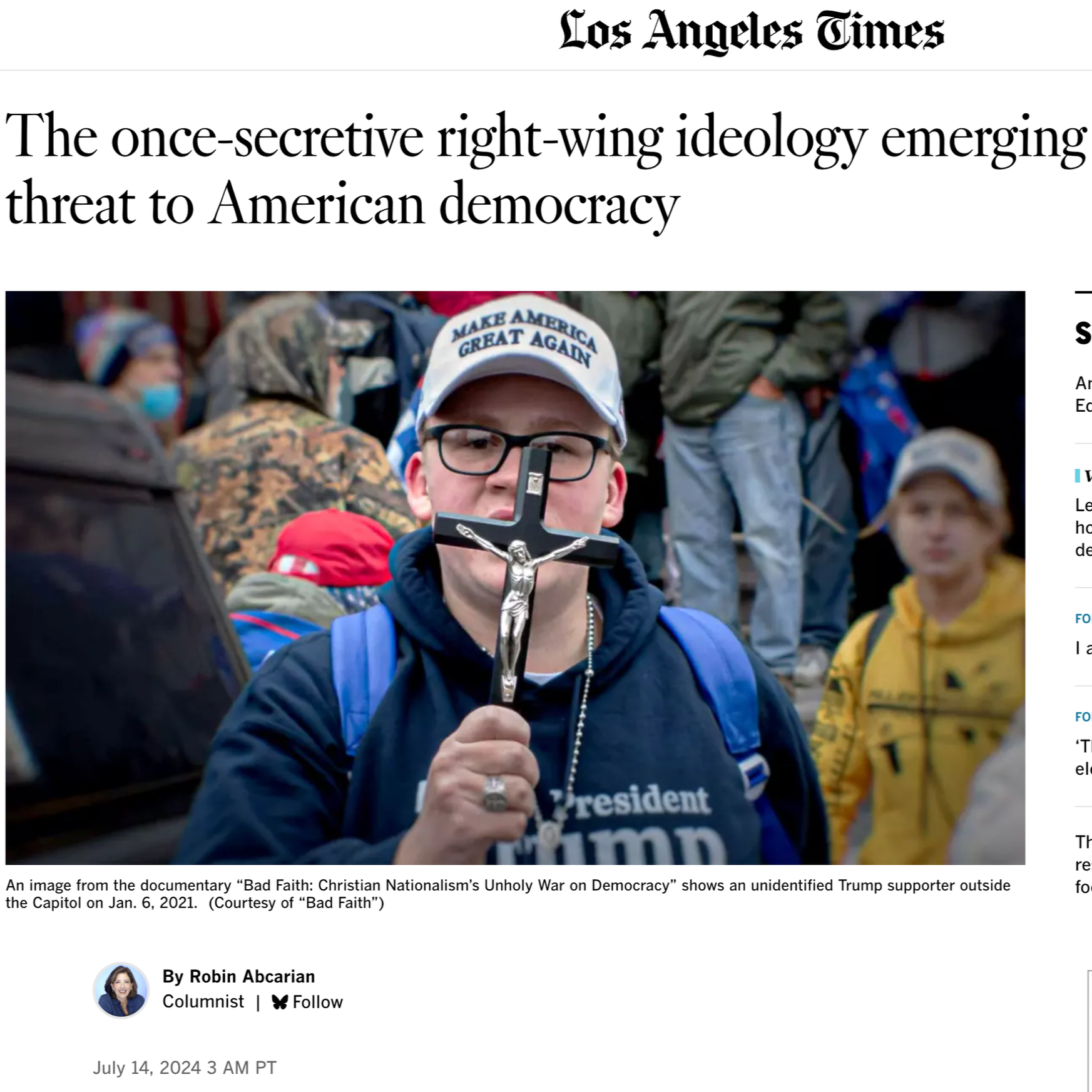 A man wearing glasses and a black sweatshirt that says "president Trump" holds a crucifix and a phone in front of his face during a rally or protest. He is surrounded by other people, some of whom are wearing masks, with one person in a yellow jacket. The scene appears to be outdoors.