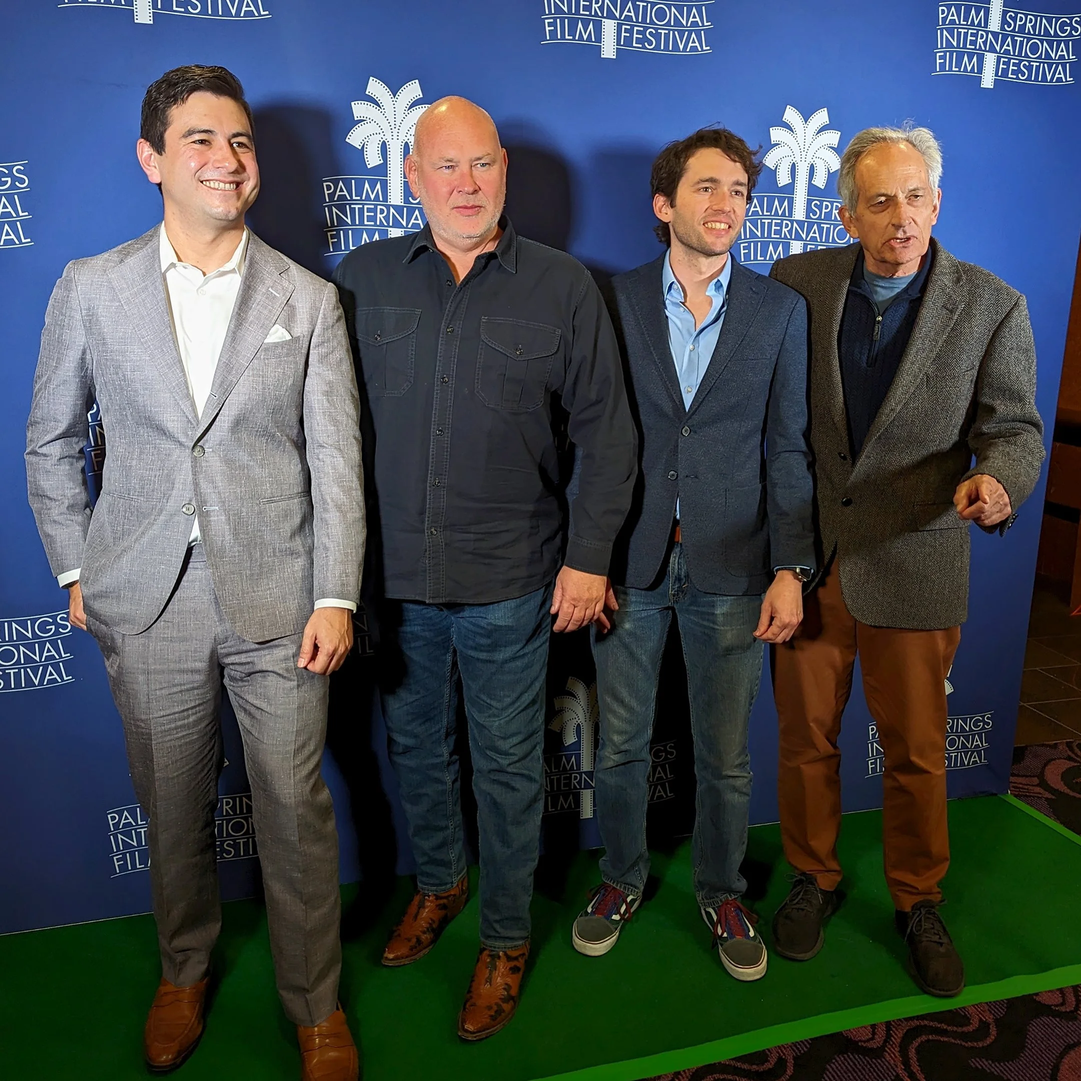 Four men are standing side by side in front of a blue backdrop with white text that reads 'Palm Springs International Film Festival.' The men are dressed in suits and casual attire, with the first man on the left wearing a light gray checked suit, the second in a dark denim jacket, the third in a dark blazer with a blue shirt, and the fourth in a plaid jacket and brown pants. They are standing on a green carpet.