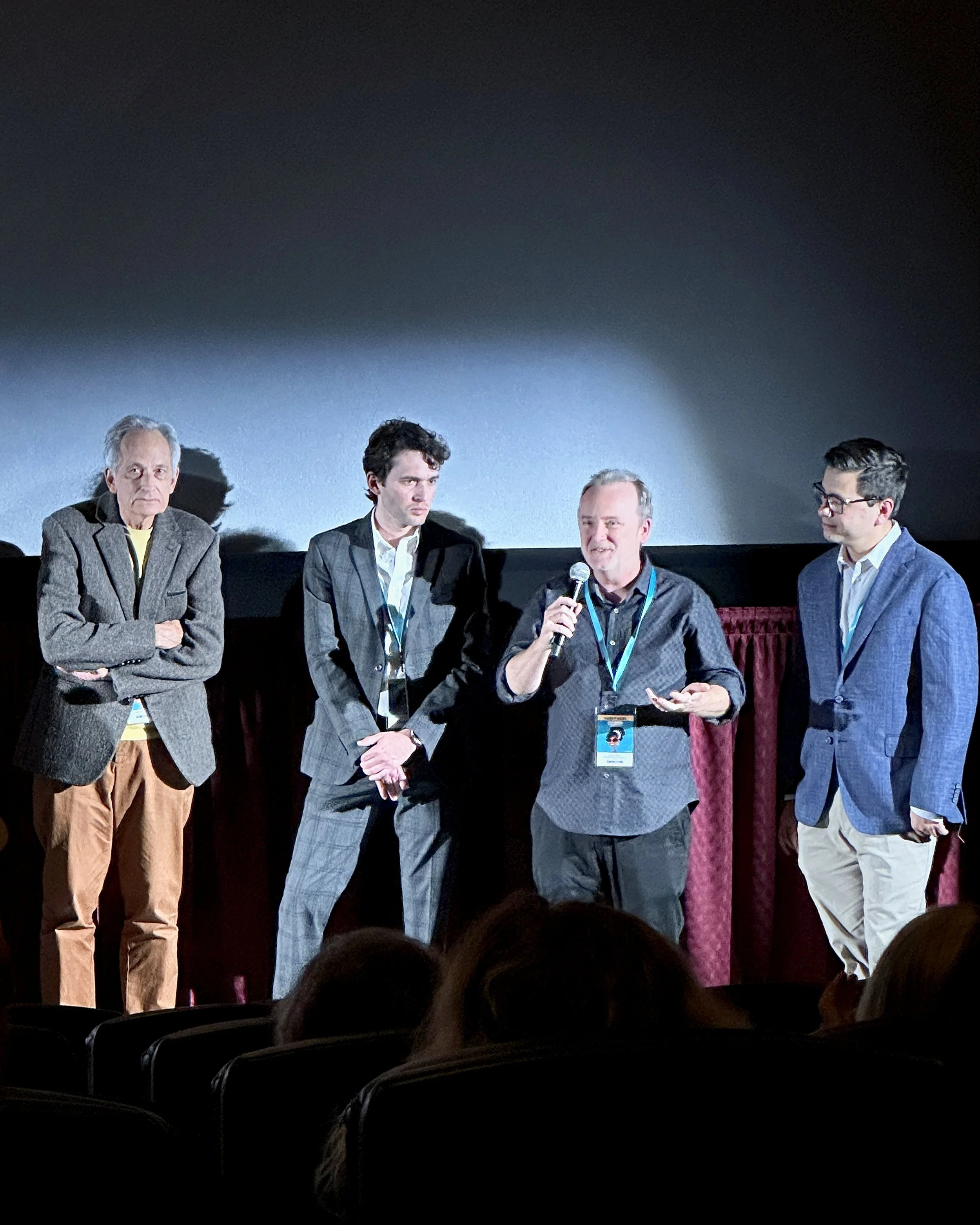 Four men standing on a stage participating in a panel discussion or presentation. Three of them are holding microphones, and one has his arms crossed.