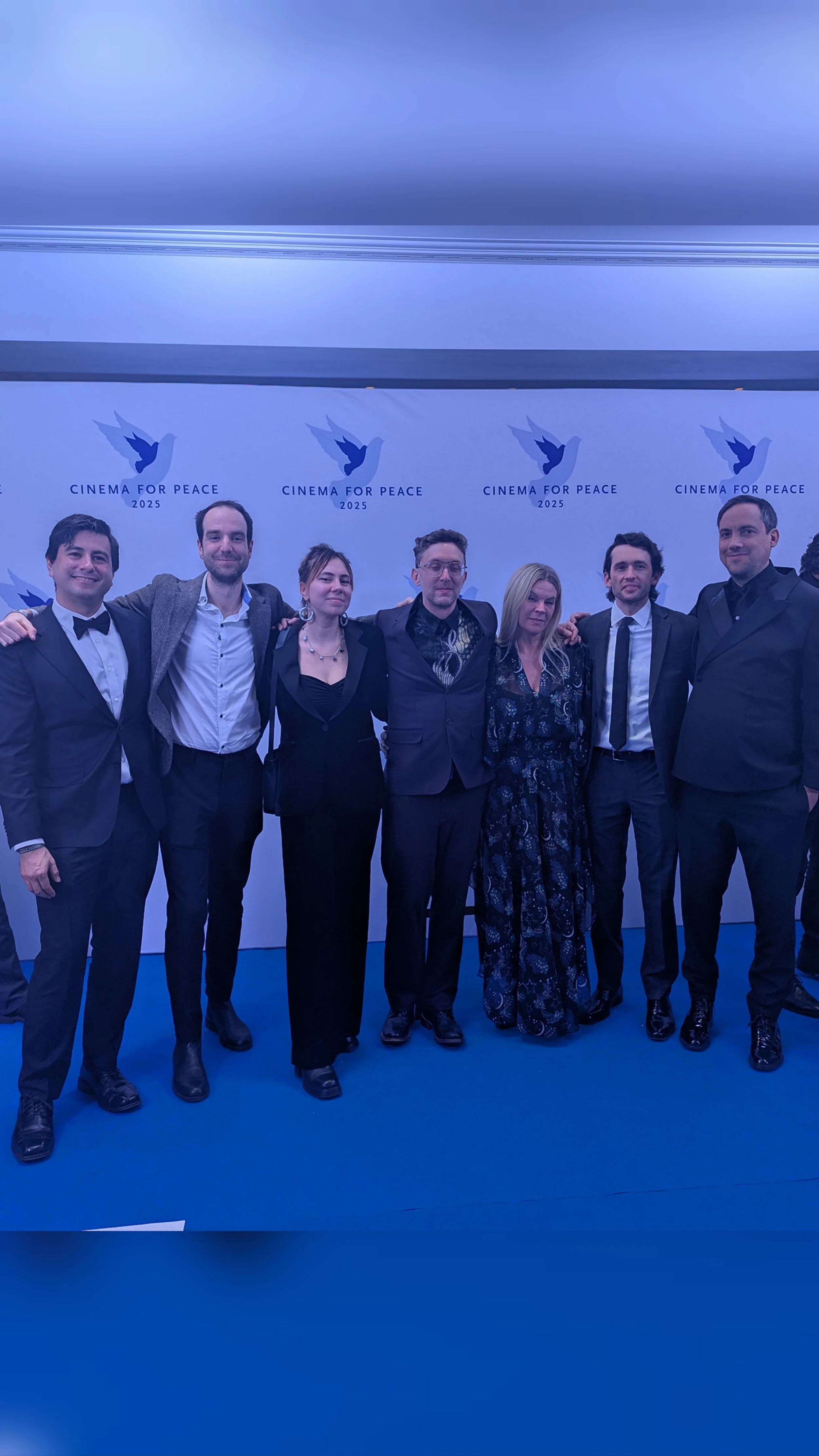 Group of seven people dressed in formal attire, standing together at the Cinema for Peace 2025 event, posing for a photo in front of a blue and white backdrop with a dove logo.