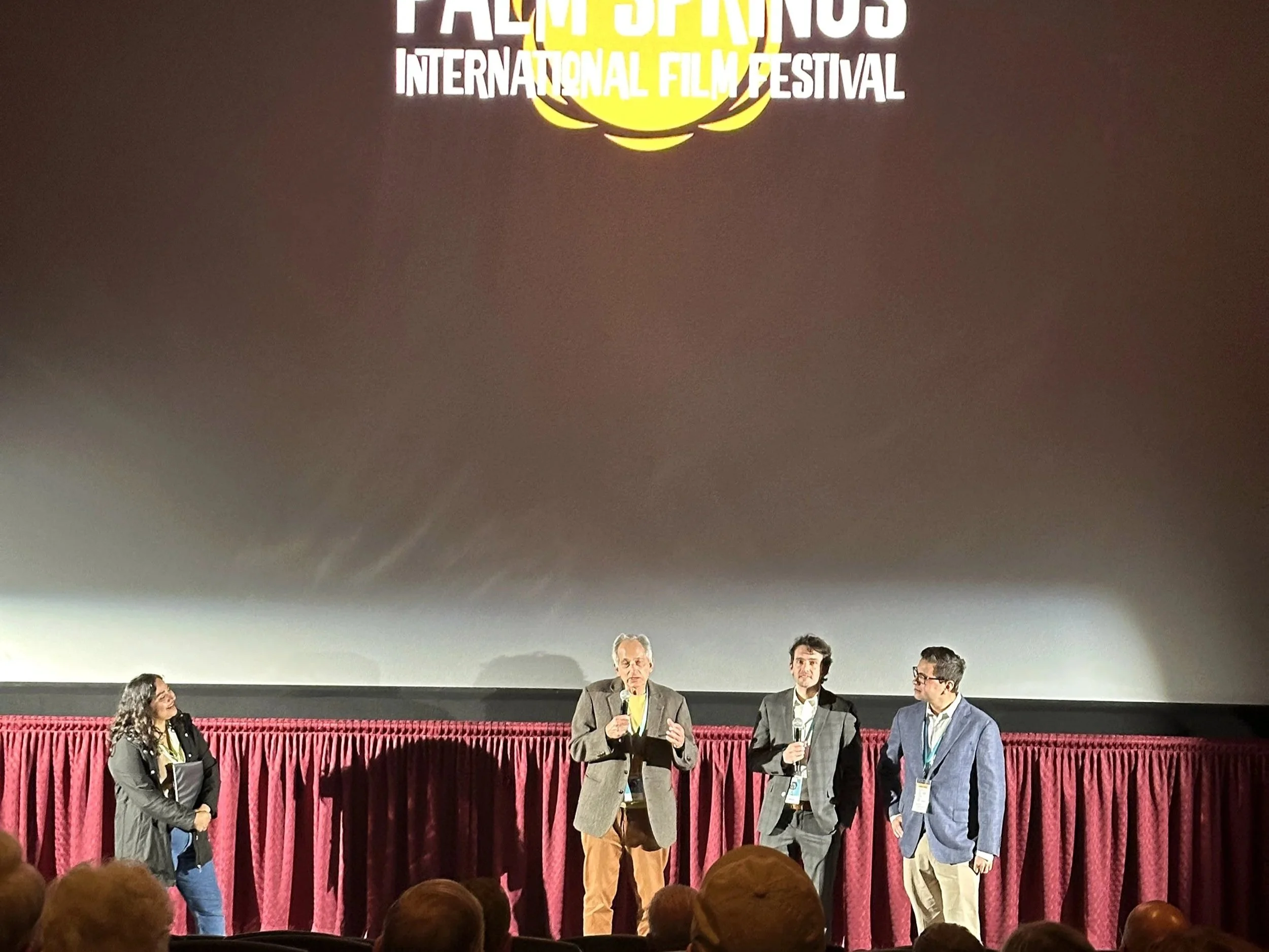 People sitting and standing on stage at the Palm Springs International Film Festival, with the festival's logo projected on a large screen behind them.