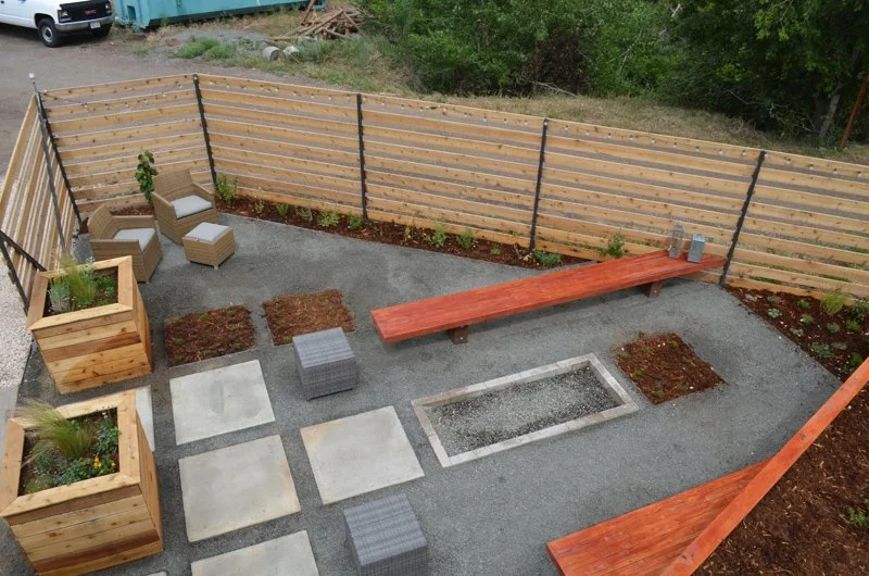 Backyard patio with wooden fencing, concrete pavers, a built-in fire pit, wooden planters with plants, a long wooden bench, and seating area with chairs.