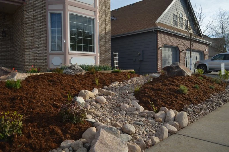 Residential landscaping with freshly tilled flower beds, decorative rocks, and a stone pathway between two houses.