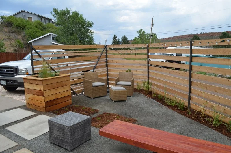 Outdoor patio with a wooden fence, two wicker chairs with cushions, a matching ottoman, a large wooden planter with a plant, and a red wooden bench, with a truck and a bus parked outside.