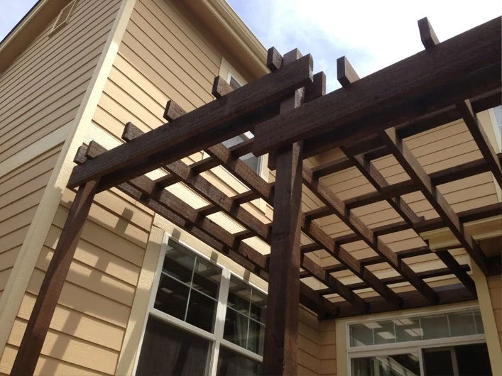 Wooden pergola structure attached to the exterior of a beige house with white trim.