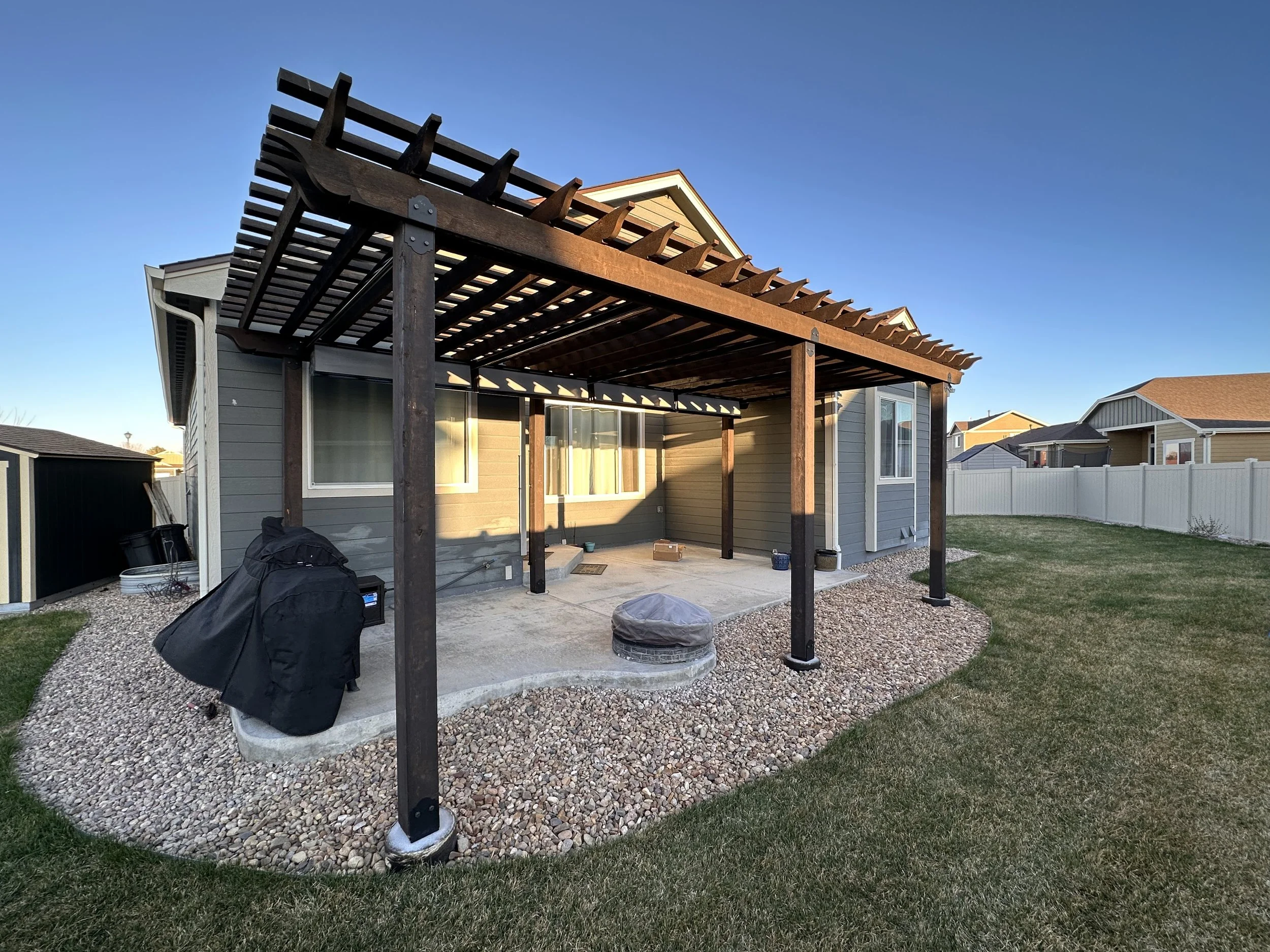 Backyard of a house with a newly built wooden pergola over concrete patio, surrounded by a white fence and green lawn.
