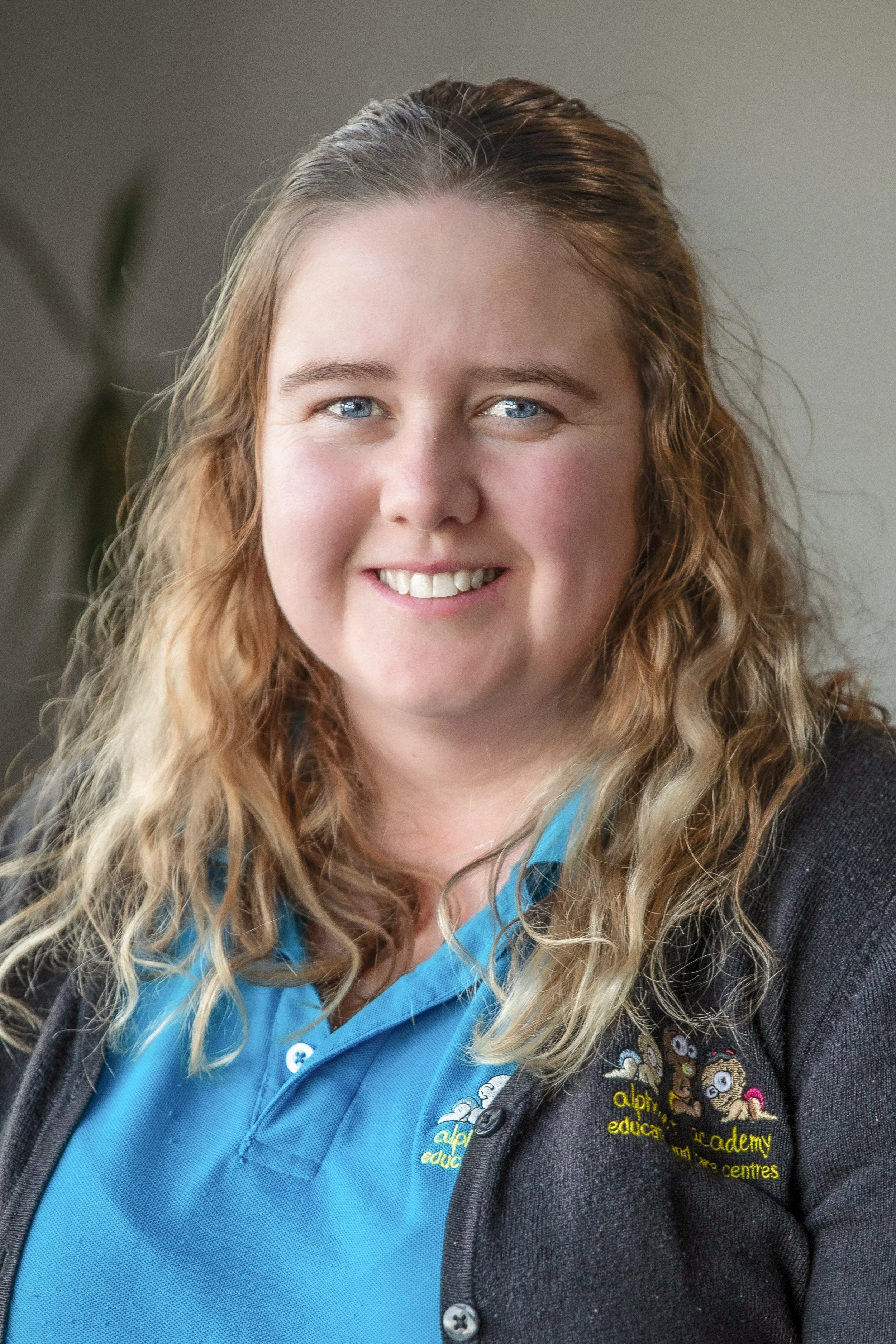 A woman with wavy, blonde hair smiling, wearing a blue polo shirt and a dark cardigan with an embroidered logo.