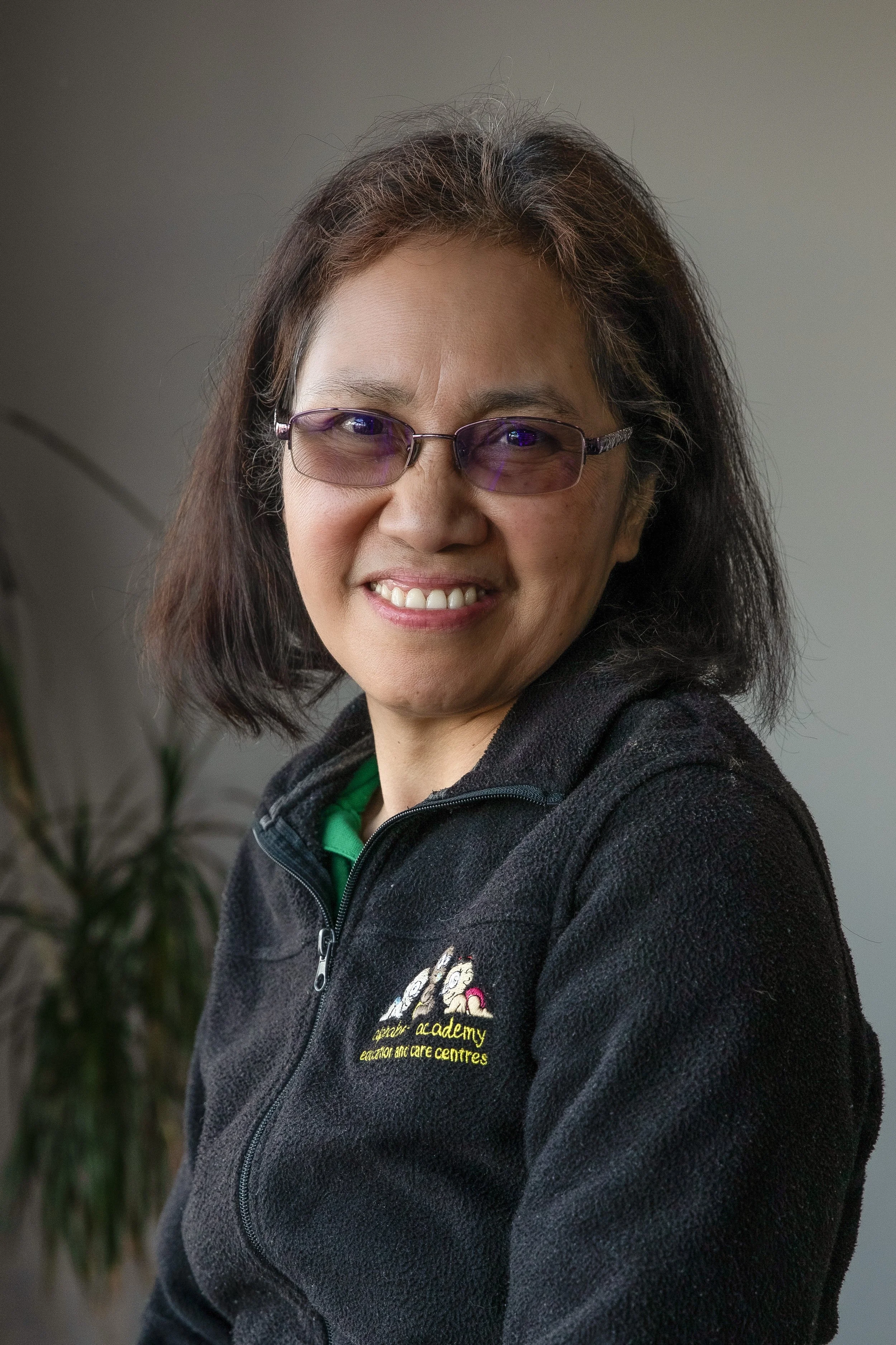 Smiling woman with glasses wearing a black fleece jacket with a logo and text on the chest, standing indoors with a plant in the background.