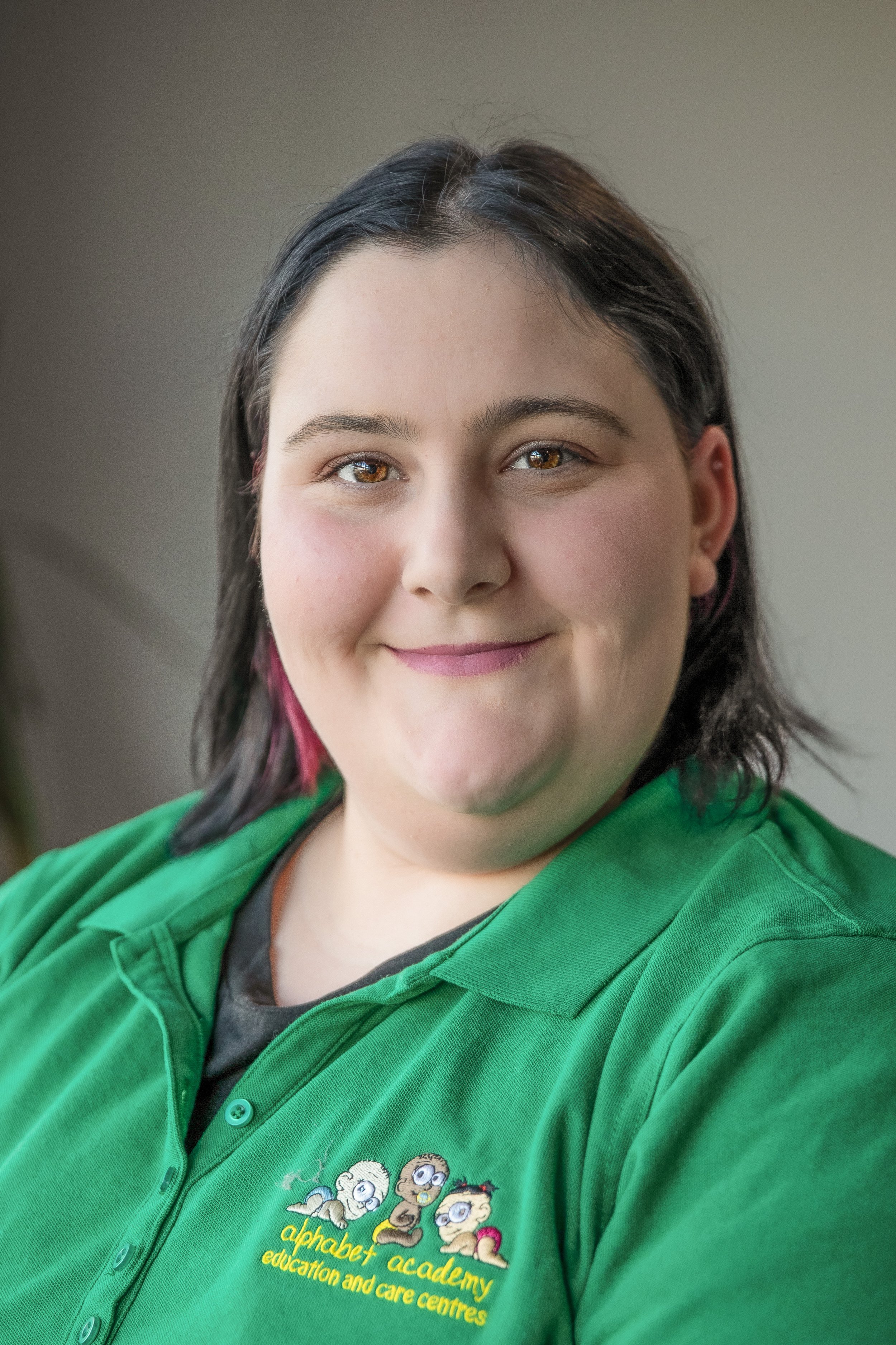 A woman with shoulder-length dark hair, brown eyes, and light skin, smiling at the camera. She is wearing a green polo shirt with a logo for AlphaBet Academy education and care centres, which features cartoon characters of children.