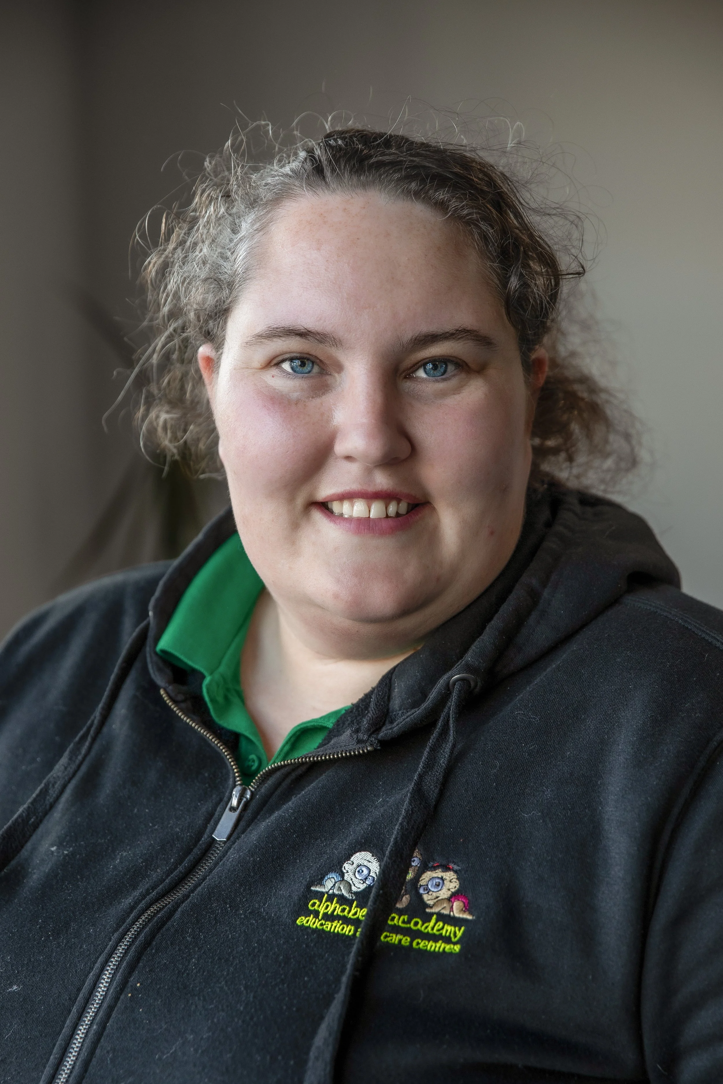 A woman with curly hair and blue eyes wearing a black jacket with a green shirt underneath, standing indoors.