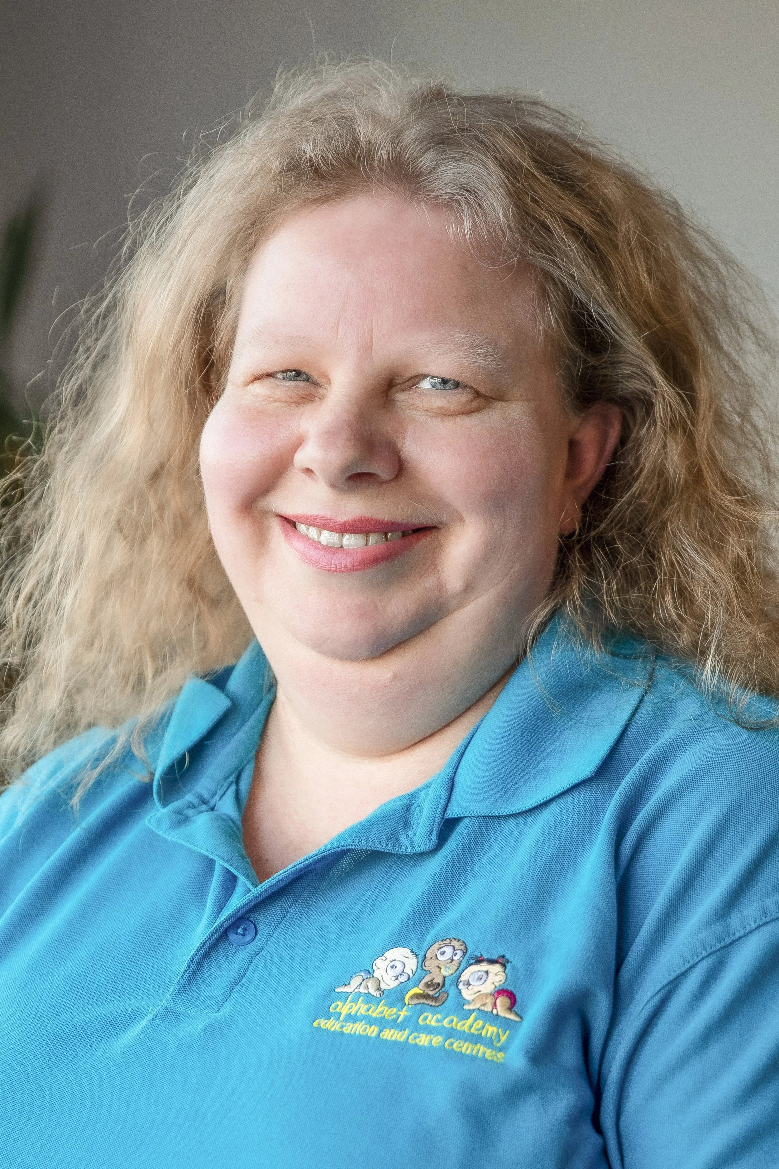 A smiling woman with curly blonde hair wearing a blue polo shirt with the logo of 'alphabet academy education and care centre' embroidered on it.