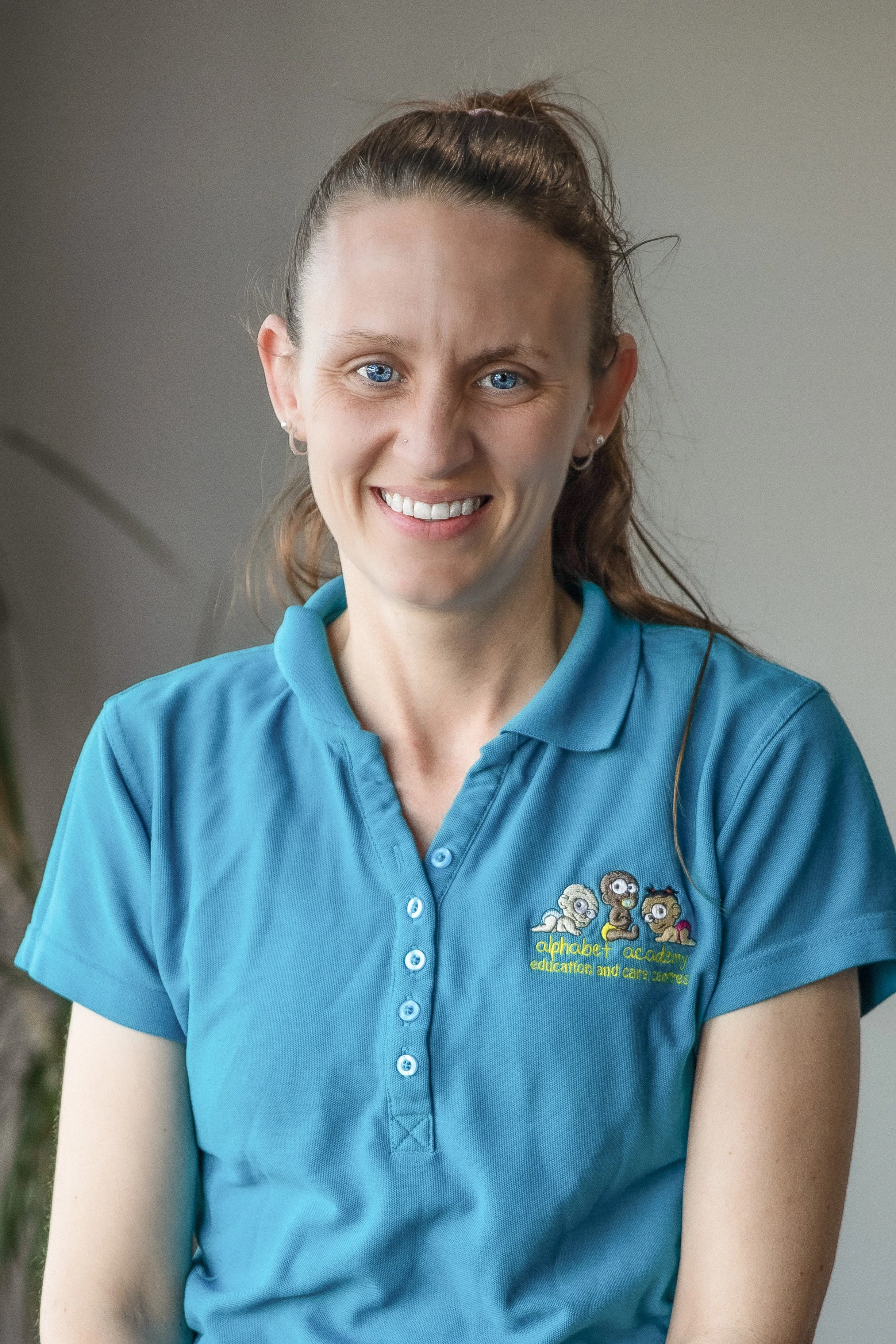 A smiling woman with blue eyes and long brown hair tied back, wearing a blue polo shirt with a logo for "alphabet academy education and care centre."