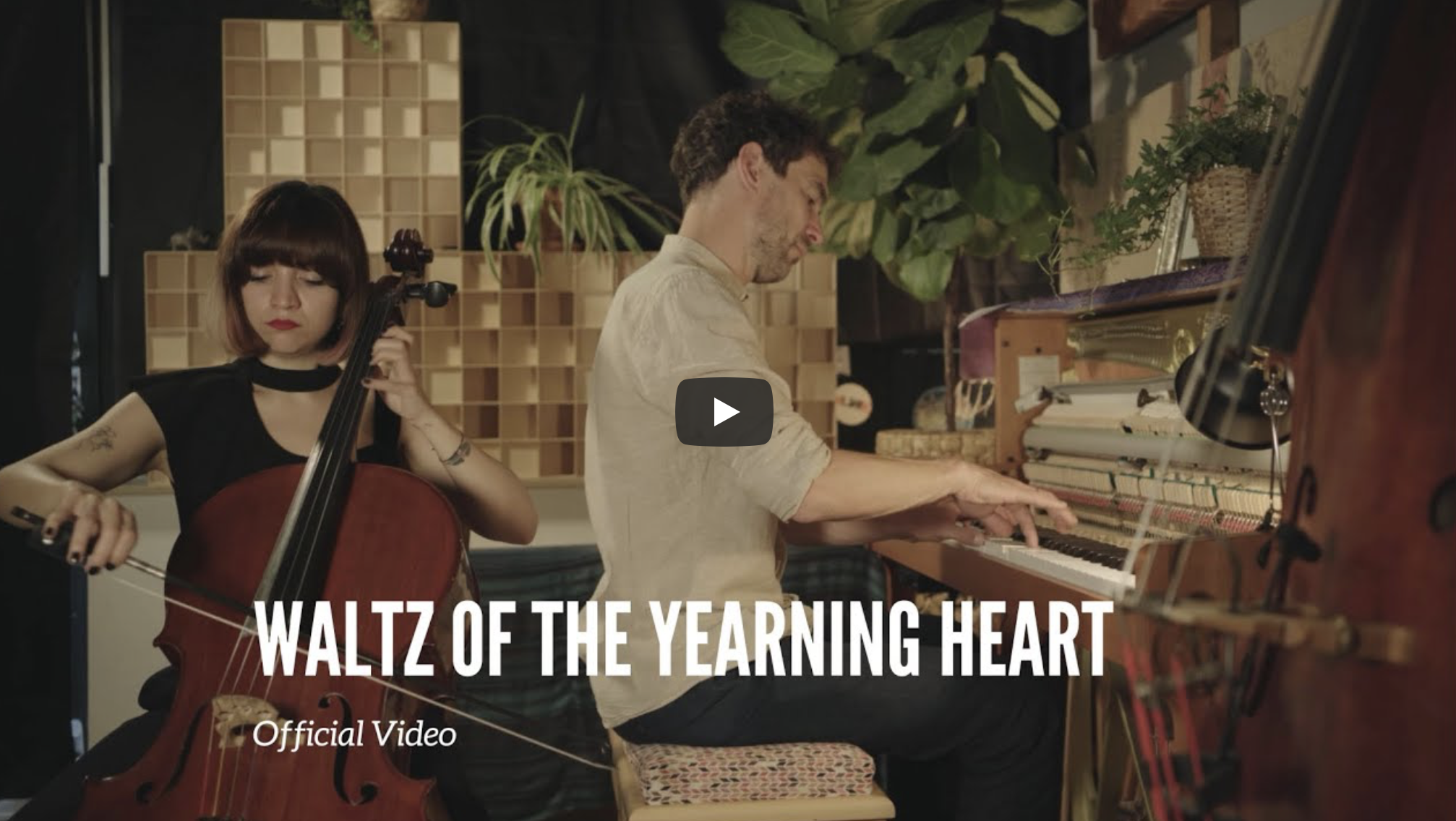 Two musicians, a woman playing a cello and a man playing a piano, in a cozy studio with plants and soundproofing panels, promoting the official music video for "Waltz of the Yearning Heart."
