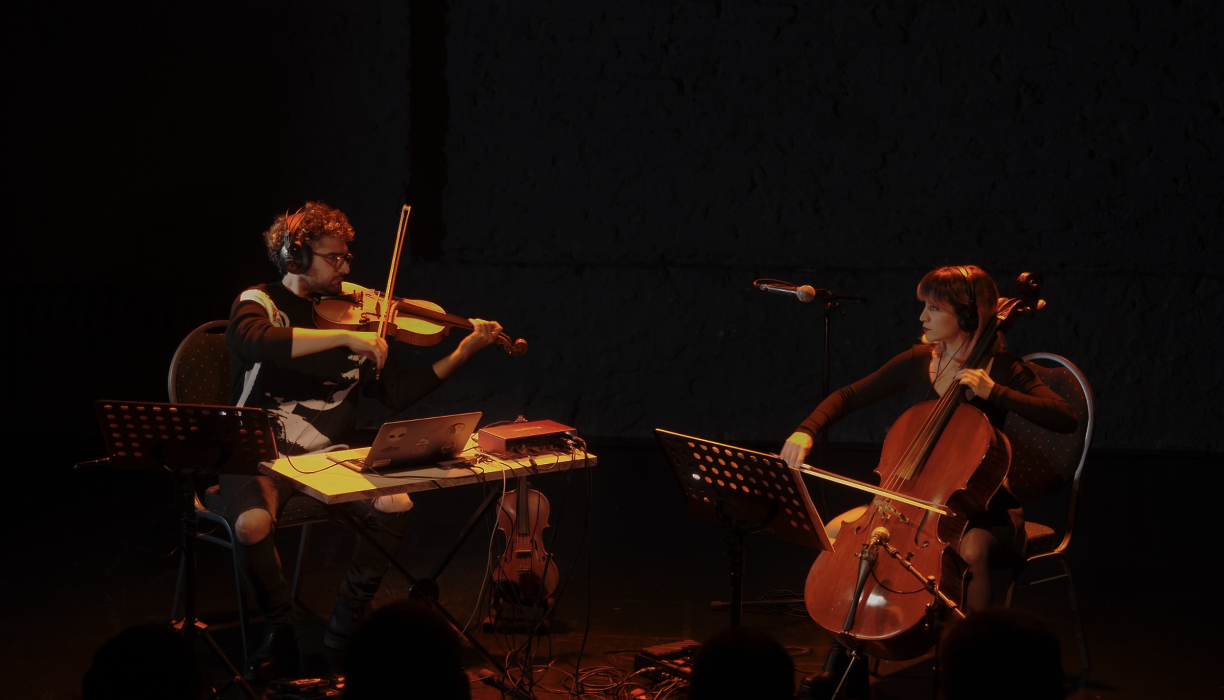 Two musicians, a man playing violin and a woman playing cello, performing on stage with music stands, chairs, and microphones in a dark, dimly lit setting.
