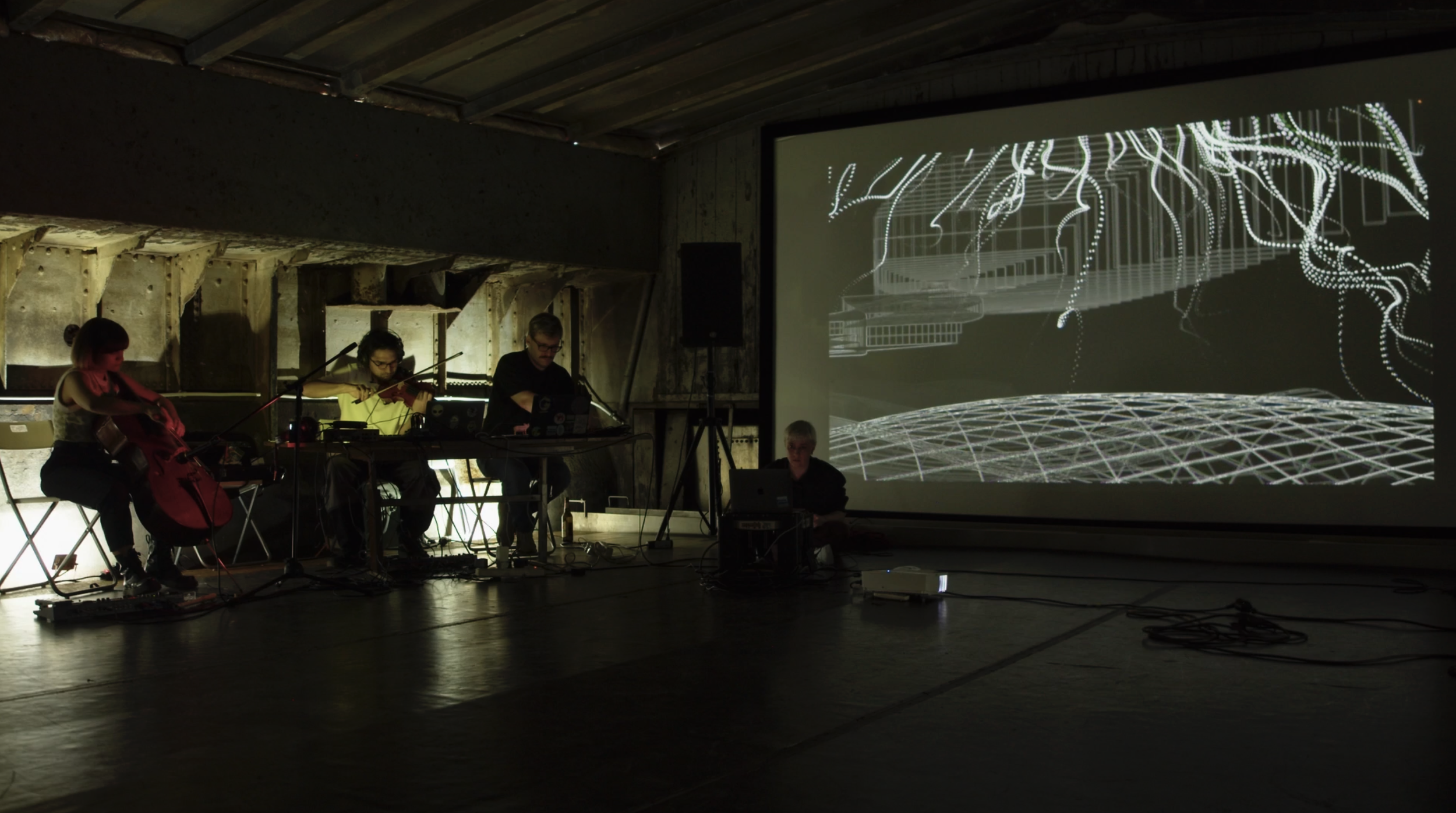 A group of four musicians performing in a dimly lit space with a large digital projection behind them displaying abstract, digital line art.