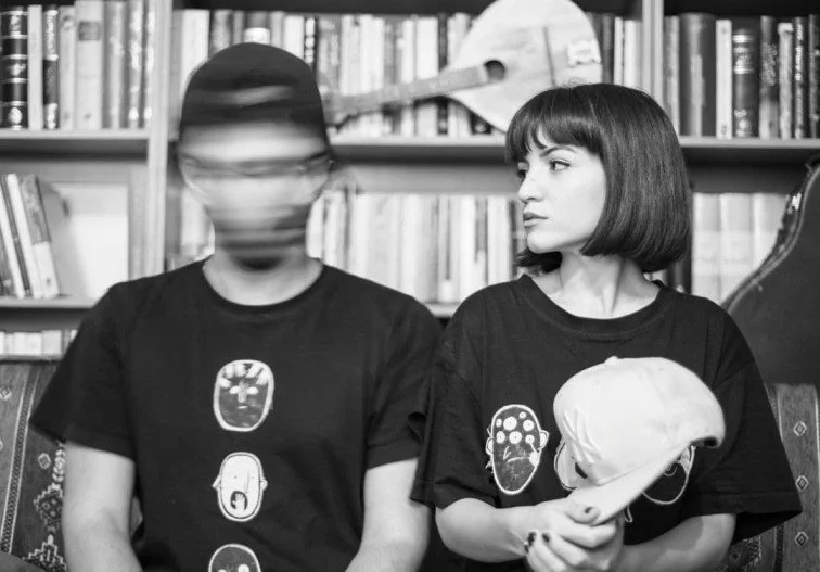 A woman with short dark hair holding a baseball cap, sitting next to a person with blurred face, both wearing black t-shirts with graphic designs, in front of a bookshelf.