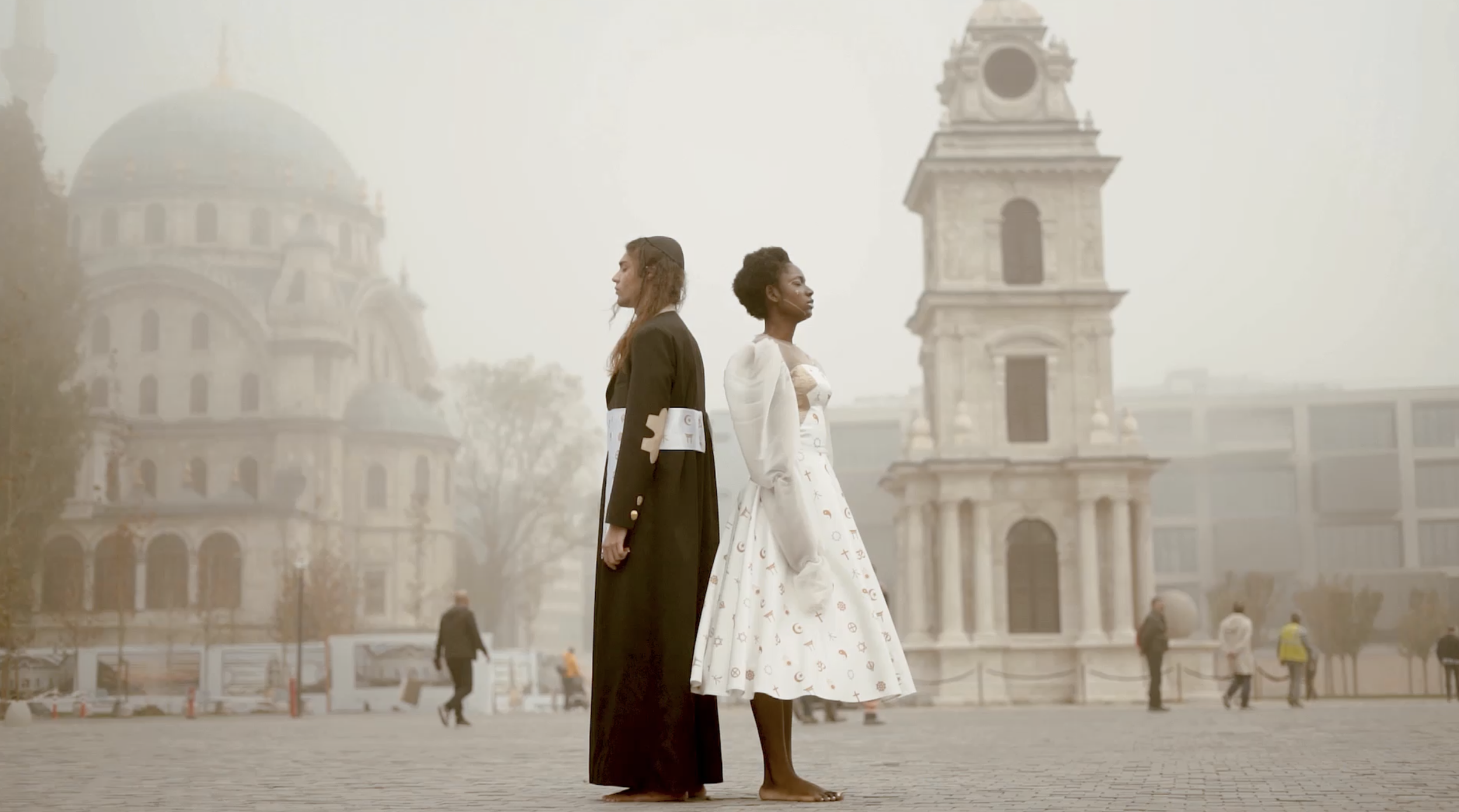 Two women stand back-to-back in a city square, with historic buildings and a clock tower in the background; they are dressed in elegant fashion, one in a black coat and the other in a white dress with symbols, both barefoot.