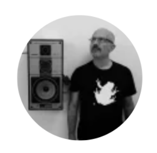 Black and white photo of a man with glasses and a beard standing next to a large speaker or sound system on the wall, in front of a plain background.