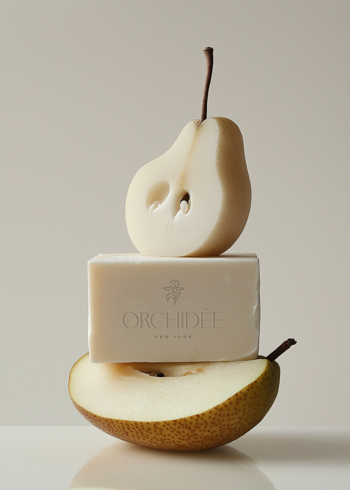 A pile of a sliced pear, a bar of soap with the word 'Orchidée' engraved on it, and another sliced pear on a flat surface with a plain light background.