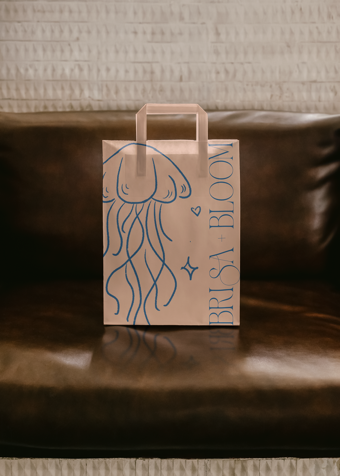 A white paper bag with a blue outline of a jellyfish and the words "FISHBRUSH & BLOOM" written vertically. The bag is placed on a brown leather surface with a textured wall in the background.