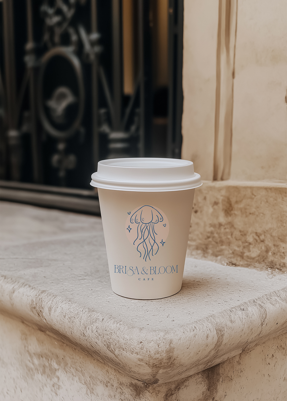 A beige paper coffee cup with a white plastic lid, featuring a blue jellyfish logo and the words "BRISA & BLOOM CAFE" on it. The cup is placed on a stone surface outdoors, with a wrought iron gate and stone wall in the background.