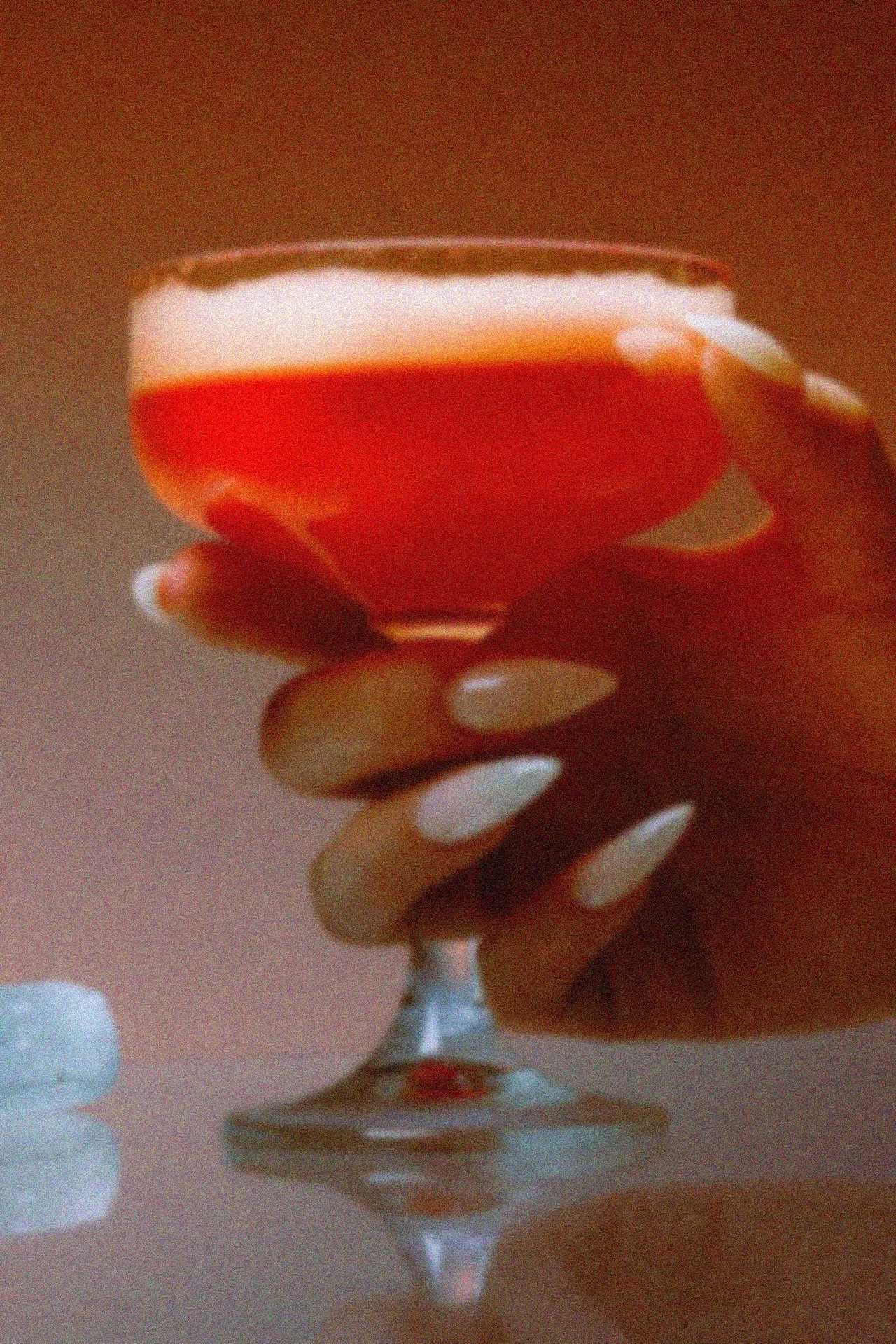 A hand holding a cocktail glass filled with an orange-colored drink topped with a pink foam layer.