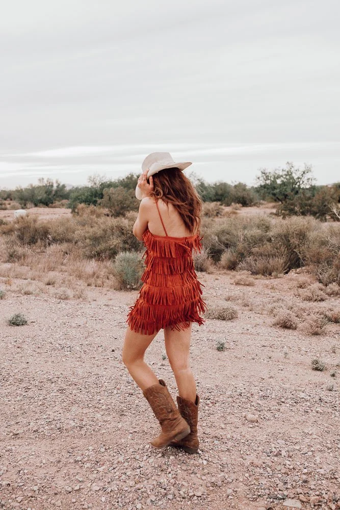 Woman in a red fringed dress, cowboy boots, and a large light-colored hat walking in a desert landscape with sparse shrubs and an overcast sky.