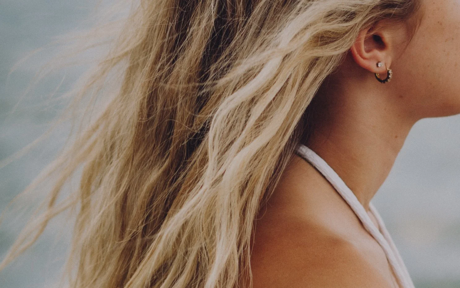 Close-up of a woman's shoulder, blonde wavy hair, and multiple earrings, with a blurred outdoor background.