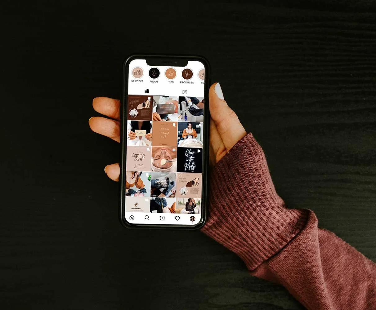 A person holding a smartphone displaying an Instagram profile with various posts, on a dark surface.
