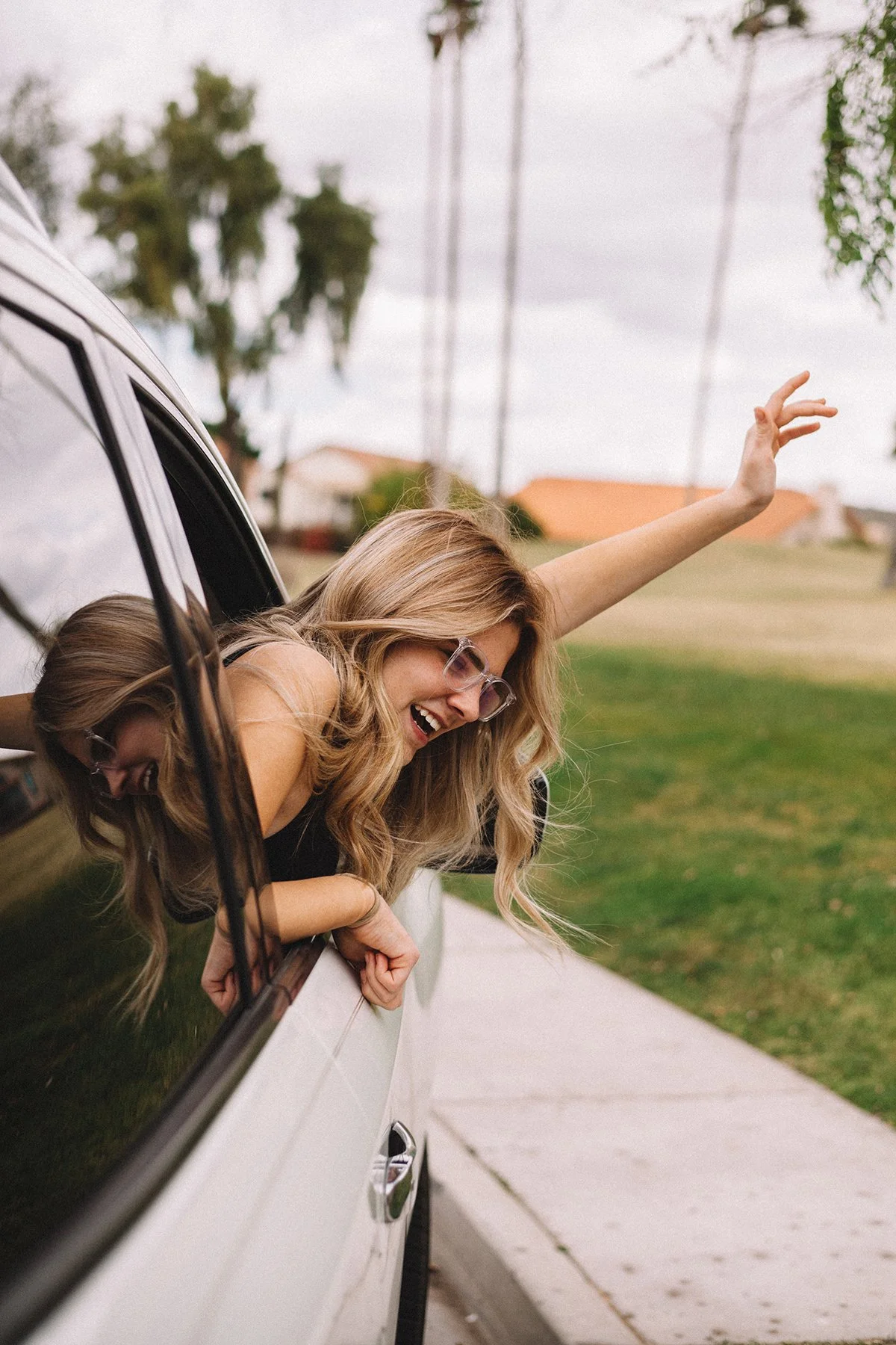 A woman with long blonde hair and sunglasses leaning out of a car window, smiling and waving, on a suburban street with grass, houses, and trees in the background.